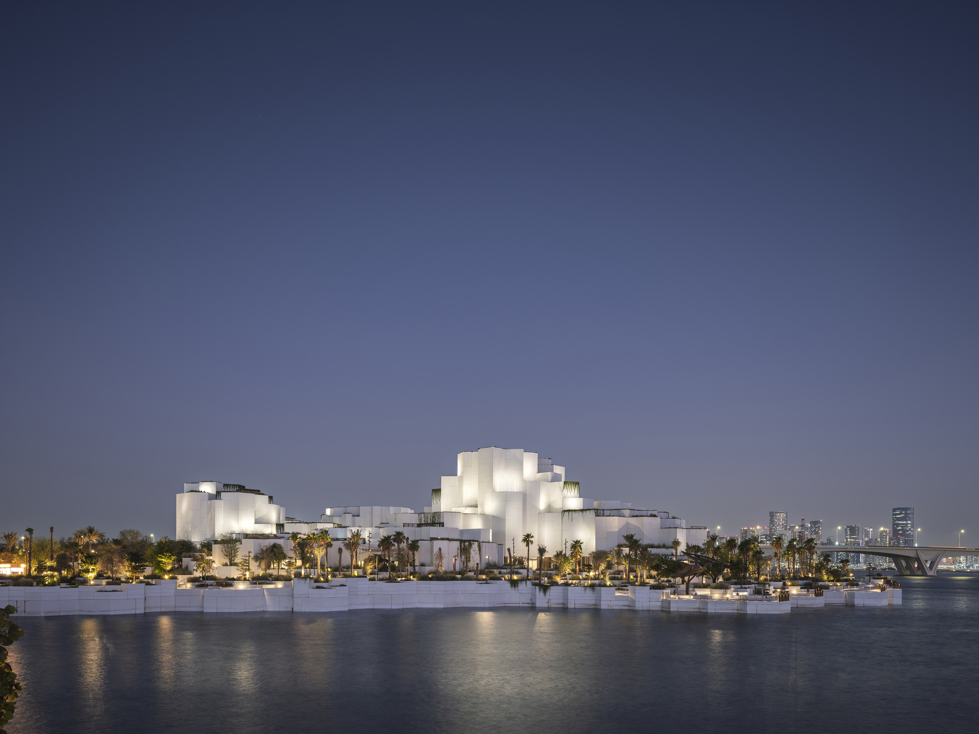 Illuminated white stepped volumes on a waterfront edge at dusk, reflected in calm water with a bridge and towers beyond