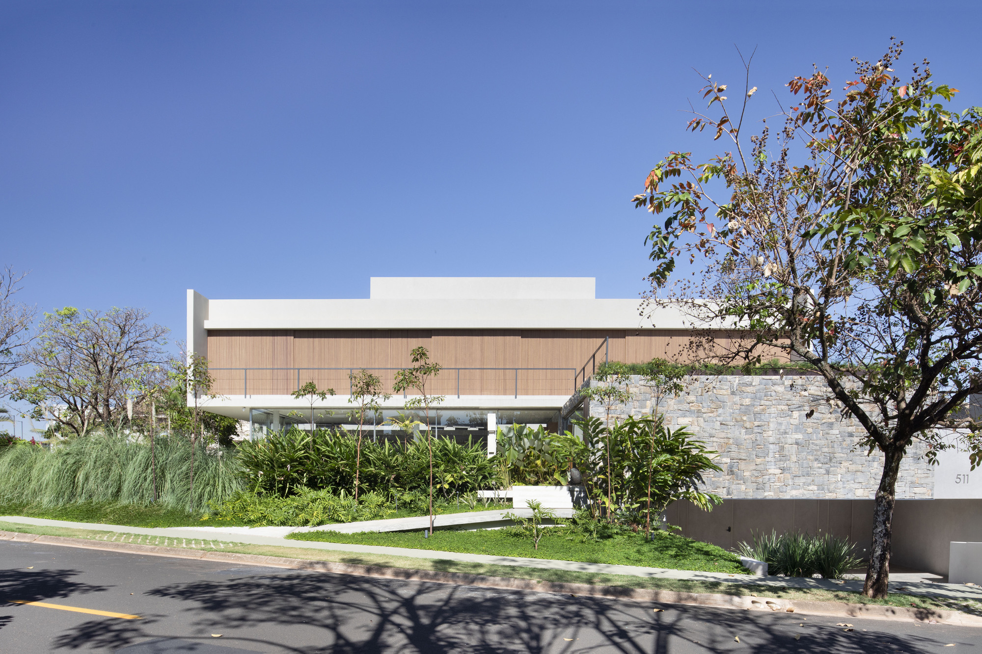 Street facade with a timber-clad upper volume above a stone retaining wall and layered tropical planting under a clear blue sky