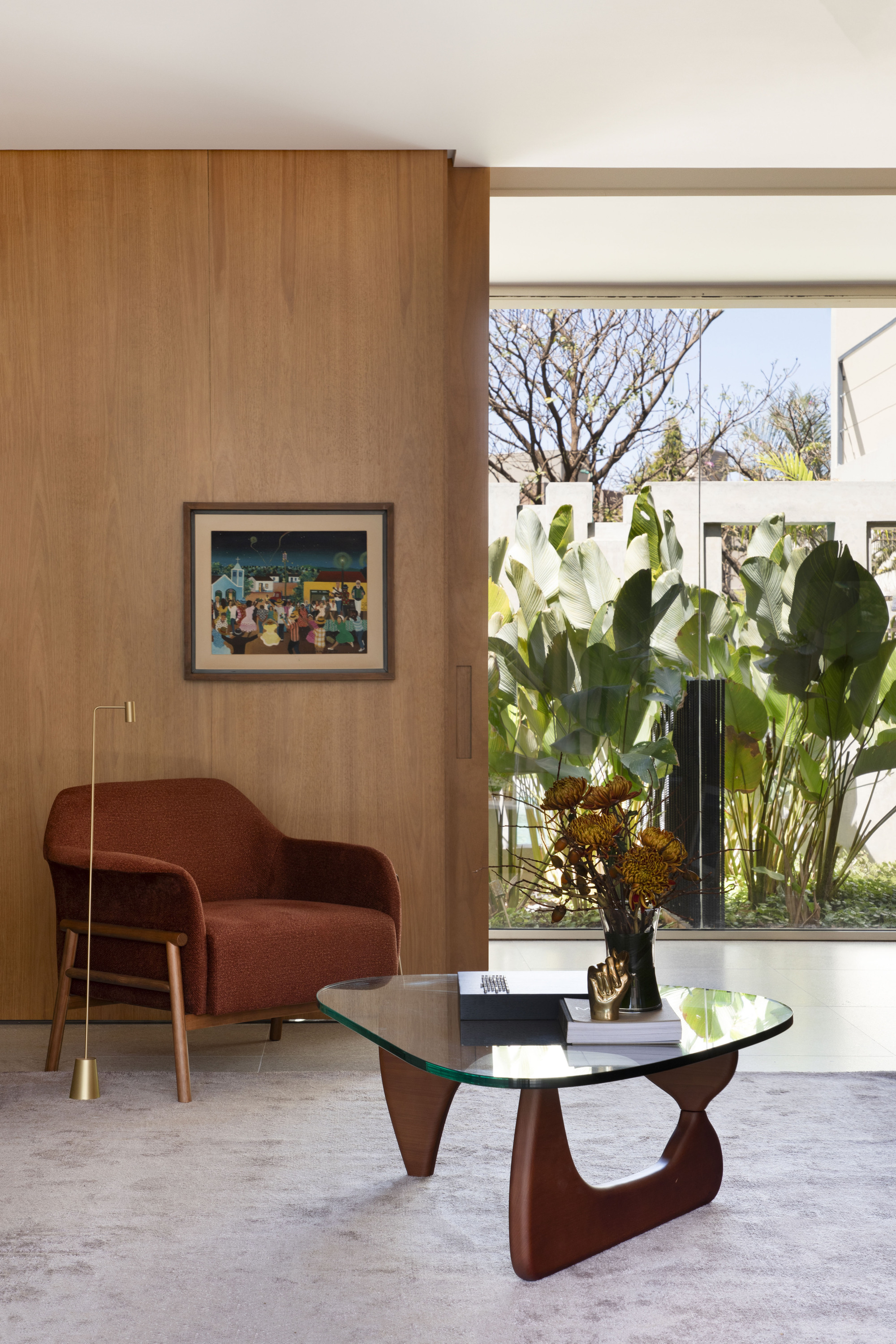 Rust-colored armchair and glass-top coffee table with dried flowers against a timber wall, banana leaves visible through the glazing