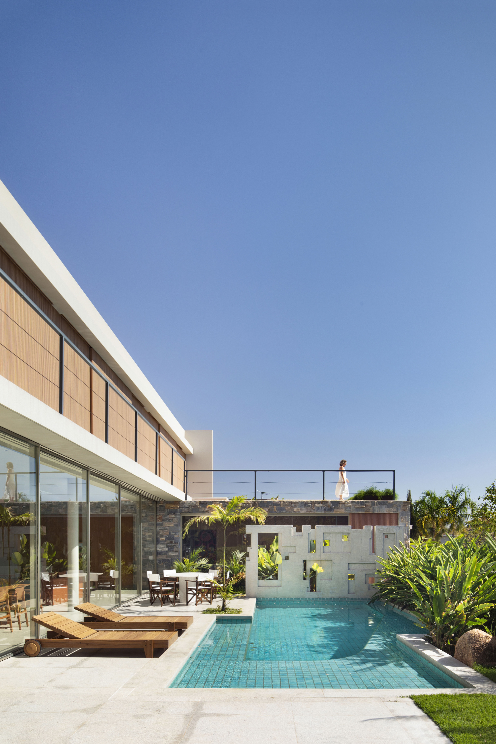 Pool terrace with teak loungers, a perforated white screen wall and a person standing on the rooftop railing in bright sun