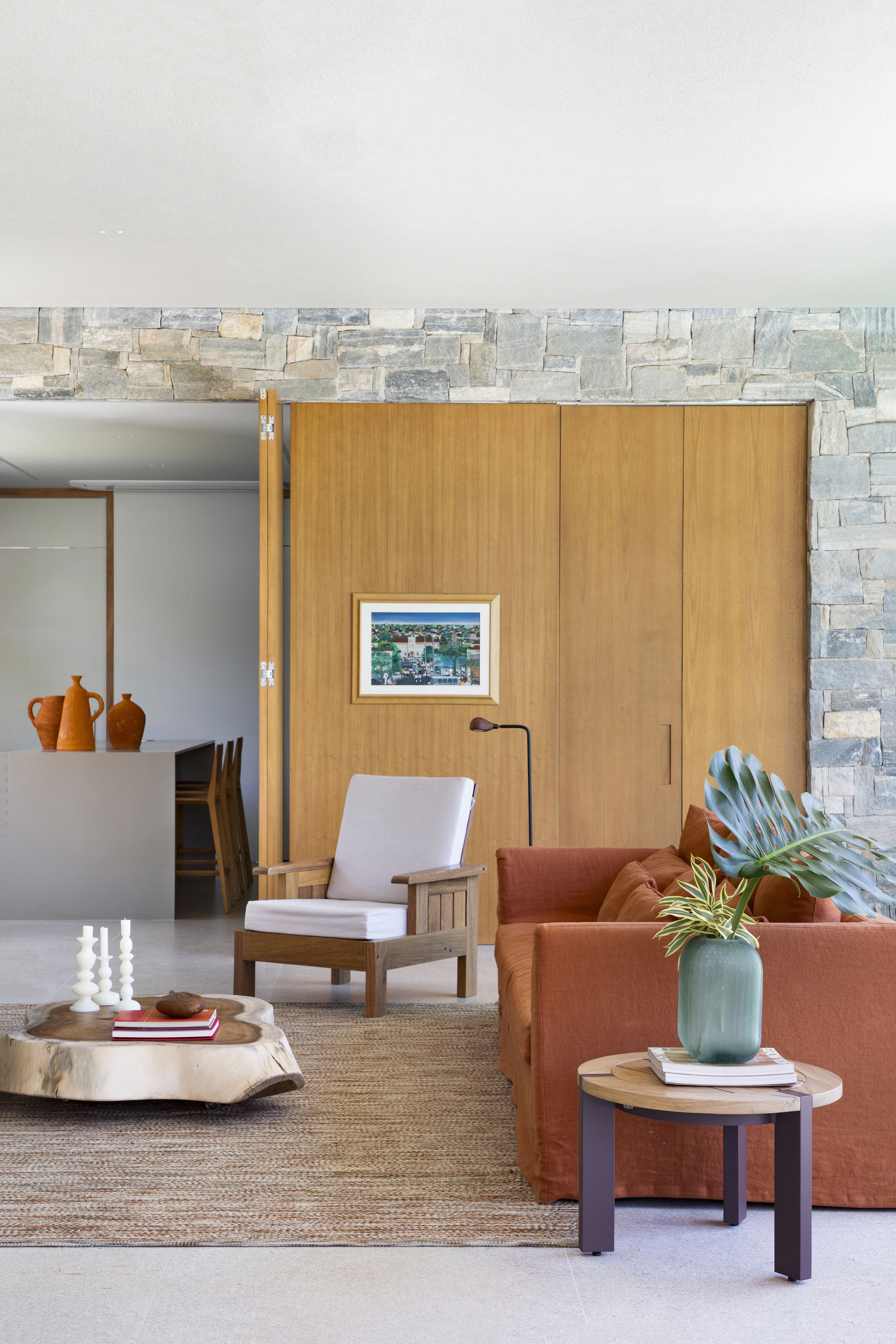 Living area with a rust linen sofa and timber armchair in front of a pivoting wood panel and rough stone wall