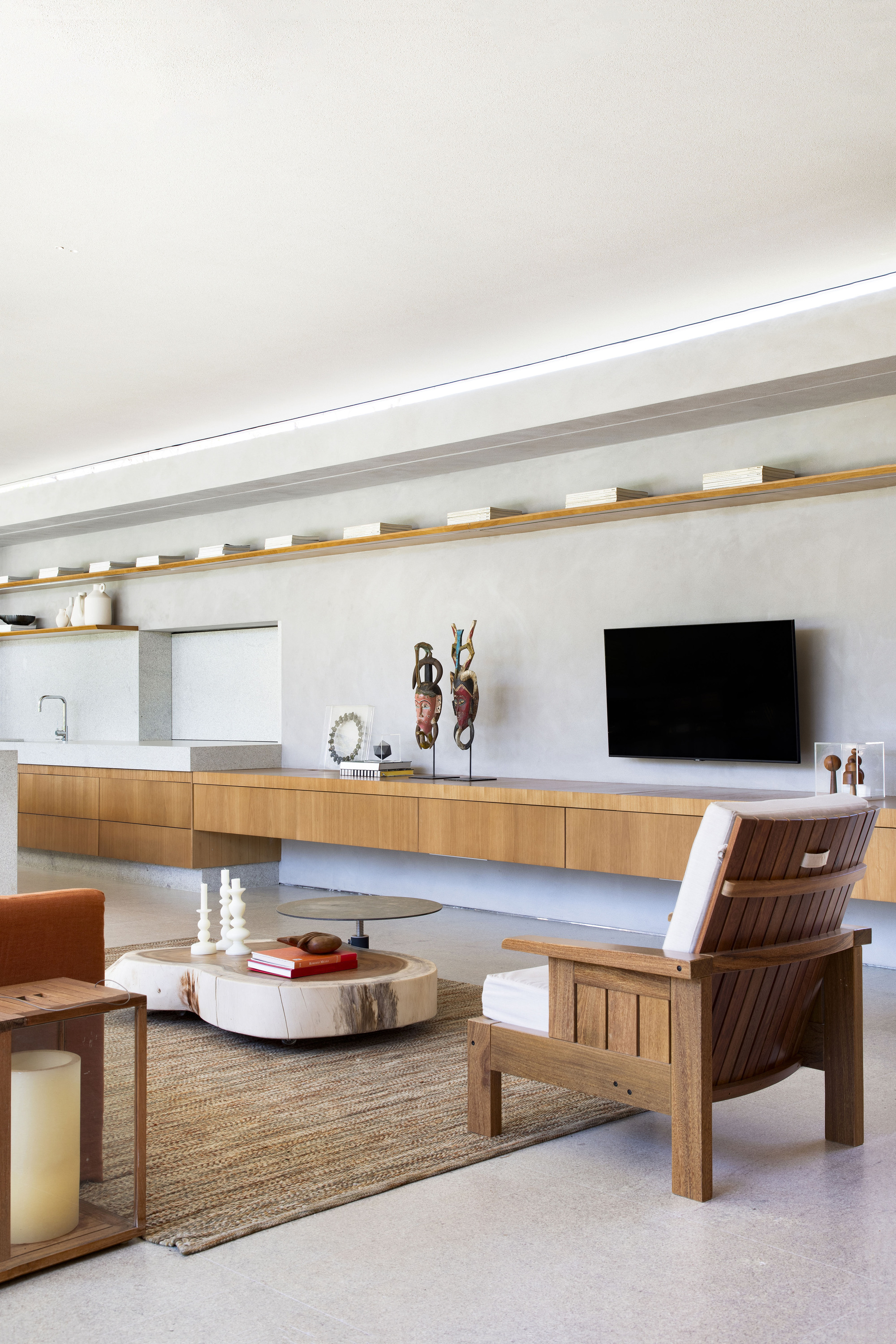 Long timber credenza and floating shelf along a plaster wall, with a slatted armchair and round coffee table in soft daylight