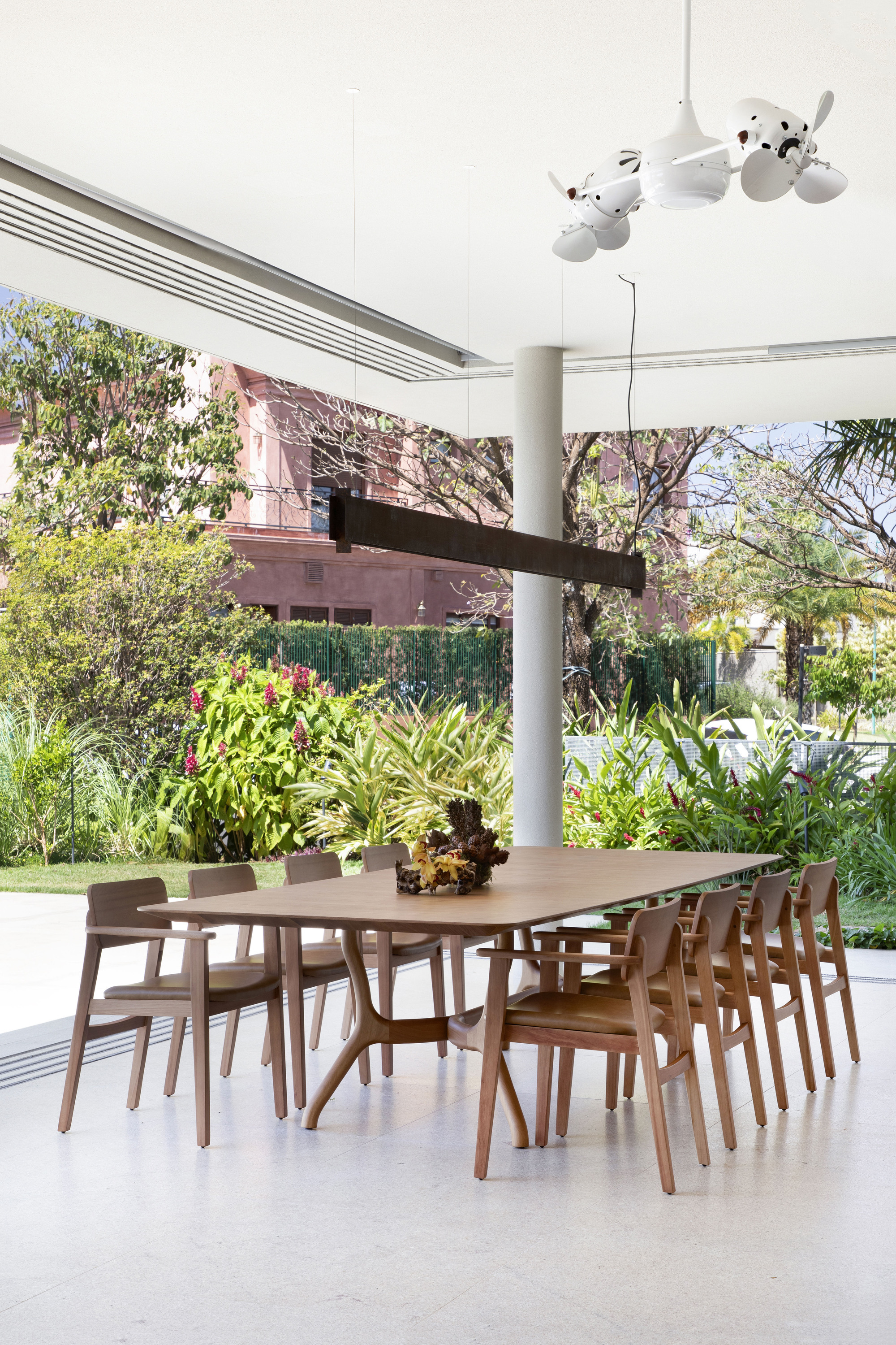 Dining table with wooden chairs beneath a white column and steel beam, open to a garden of flowering shrubs