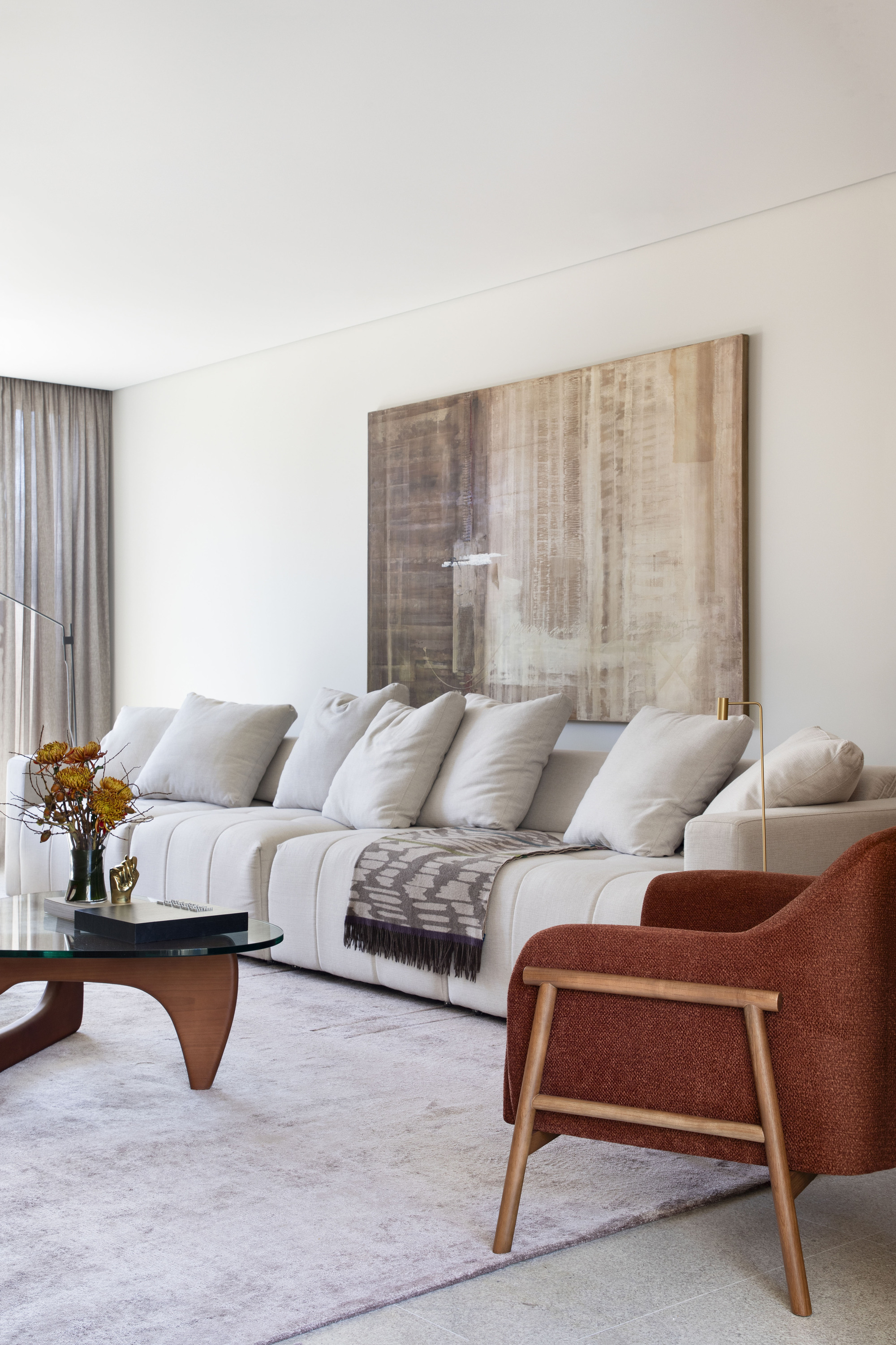 Cream sectional sofa with cushions below a large abstract canvas, with a rust armchair and glass-top table beside sheer curtains