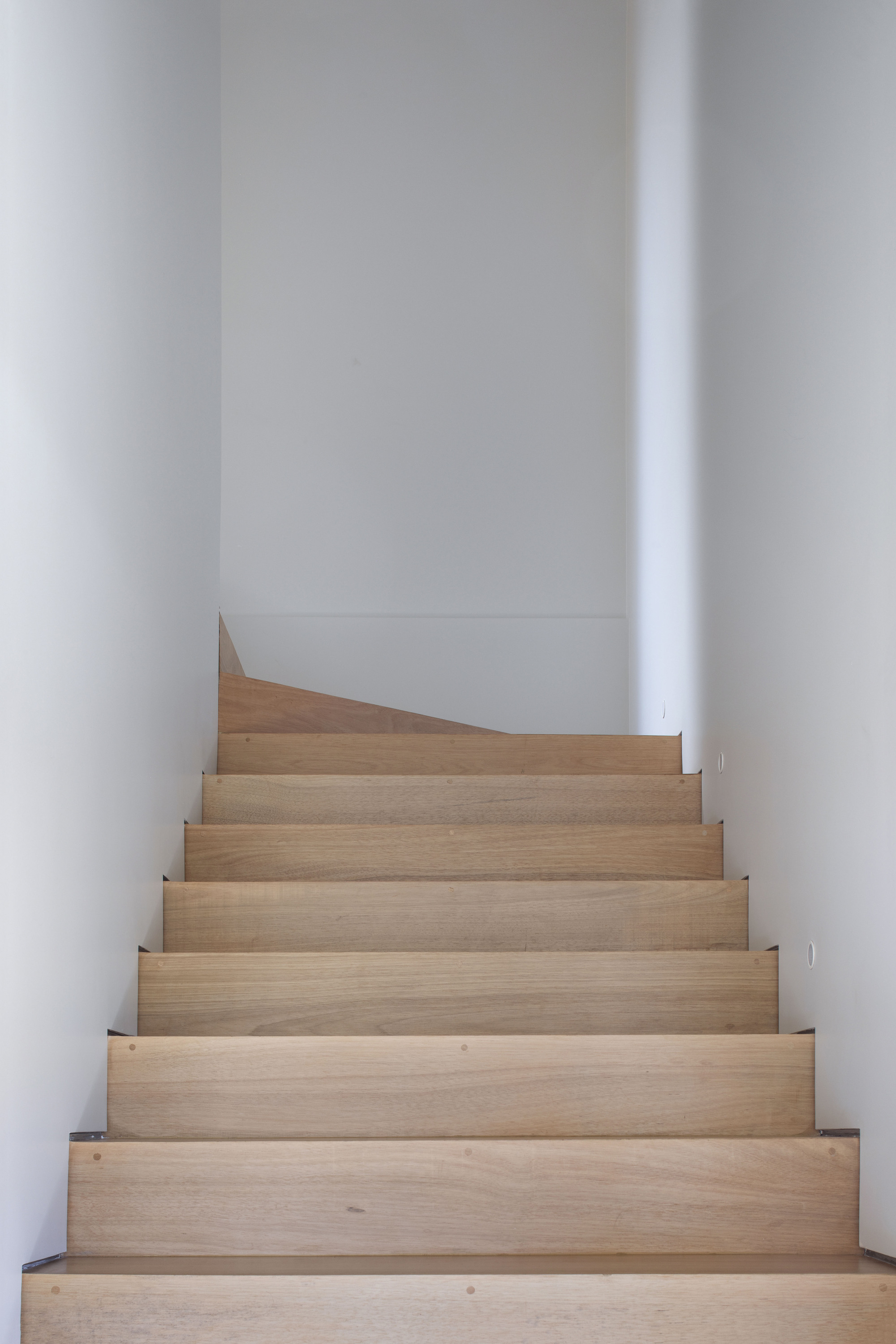 Narrow timber staircase rising between white walls to a landing with an angled wall edge in soft daylight