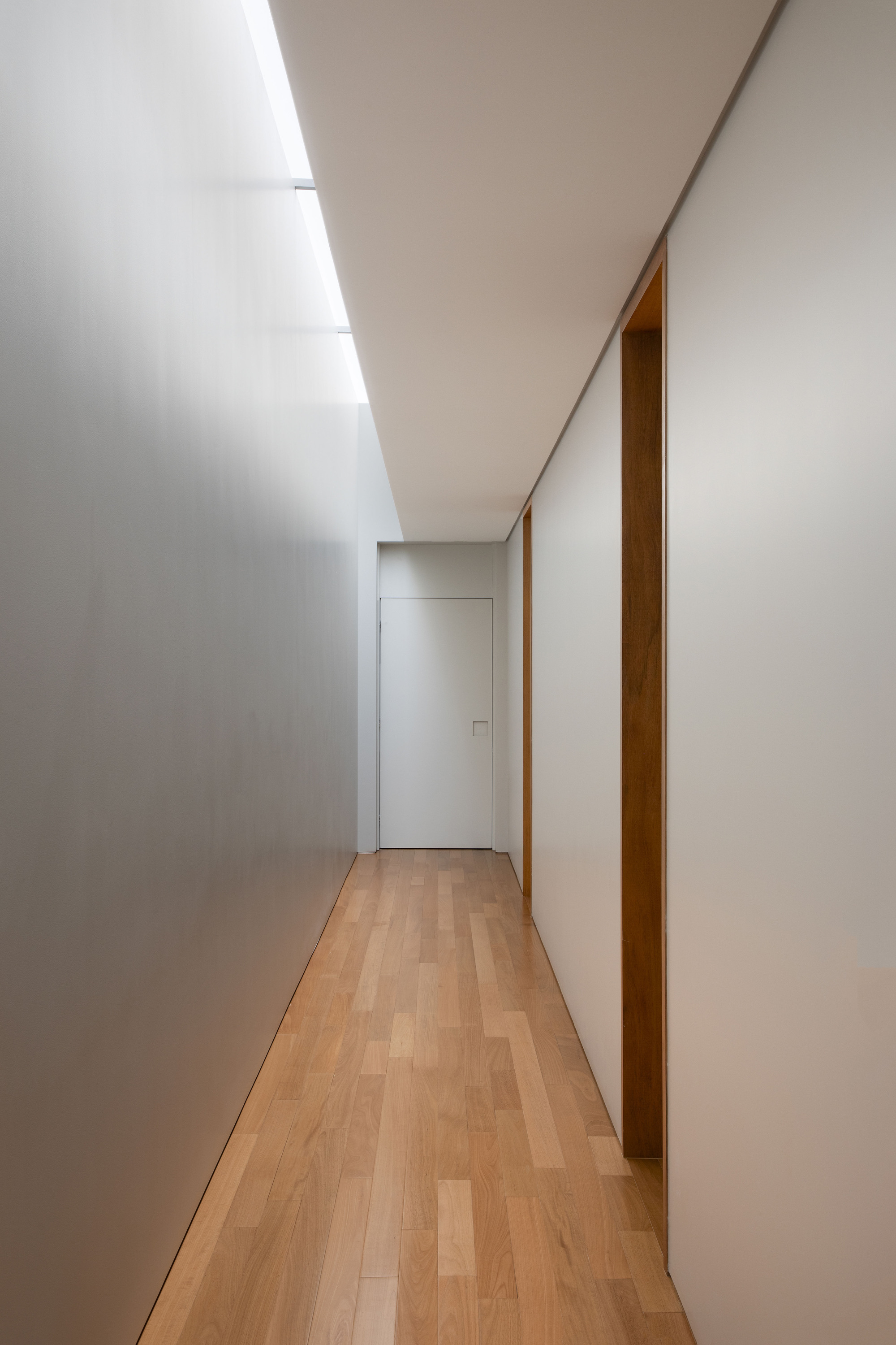 Long corridor with oak floorboards, a white flush door at the end and a skylight slot washing the left wall