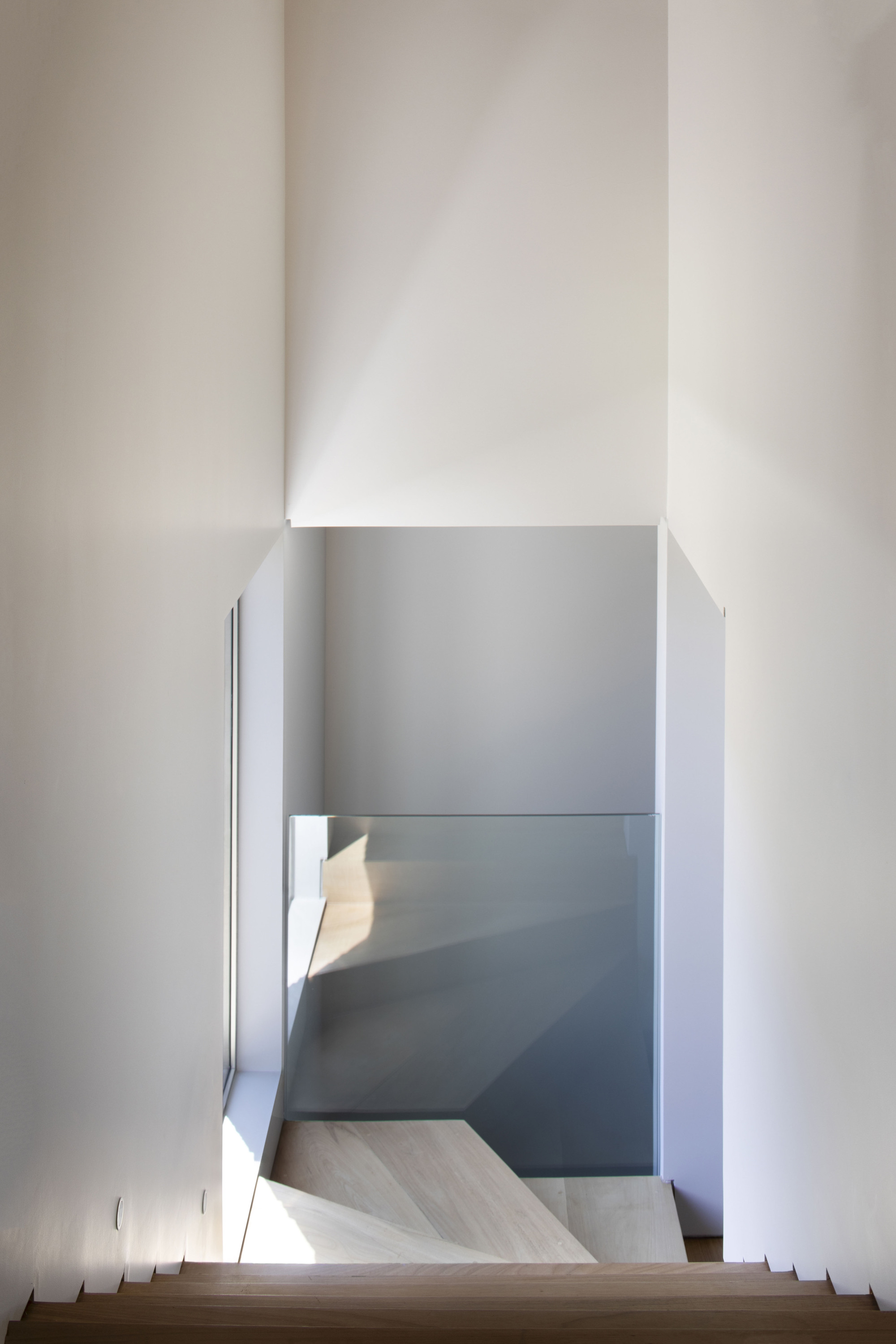 View down a staircase to a glass balustrade and pale timber landing, with sunlight cast across the walls