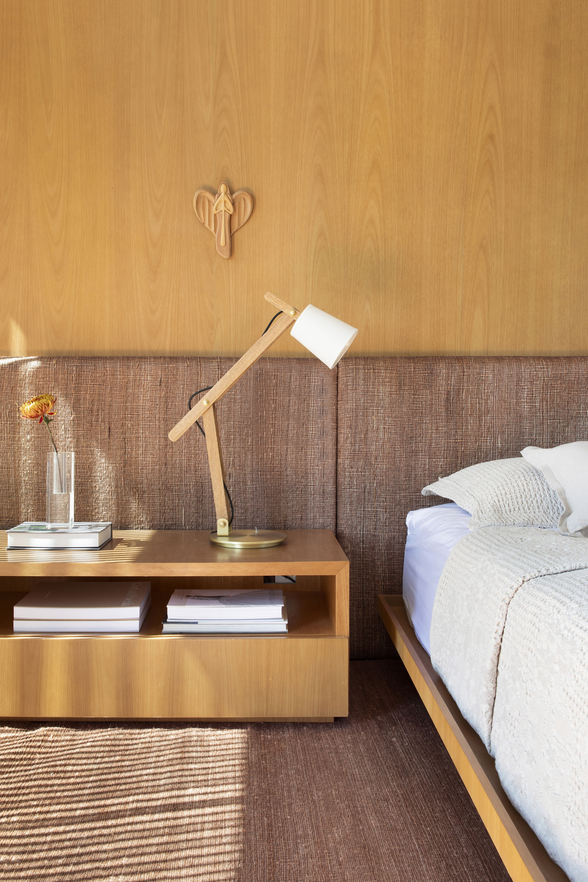 Bedside detail with oak nightstand, adjustable wooden lamp and woven wall panel, striped sunlight falling across the rug