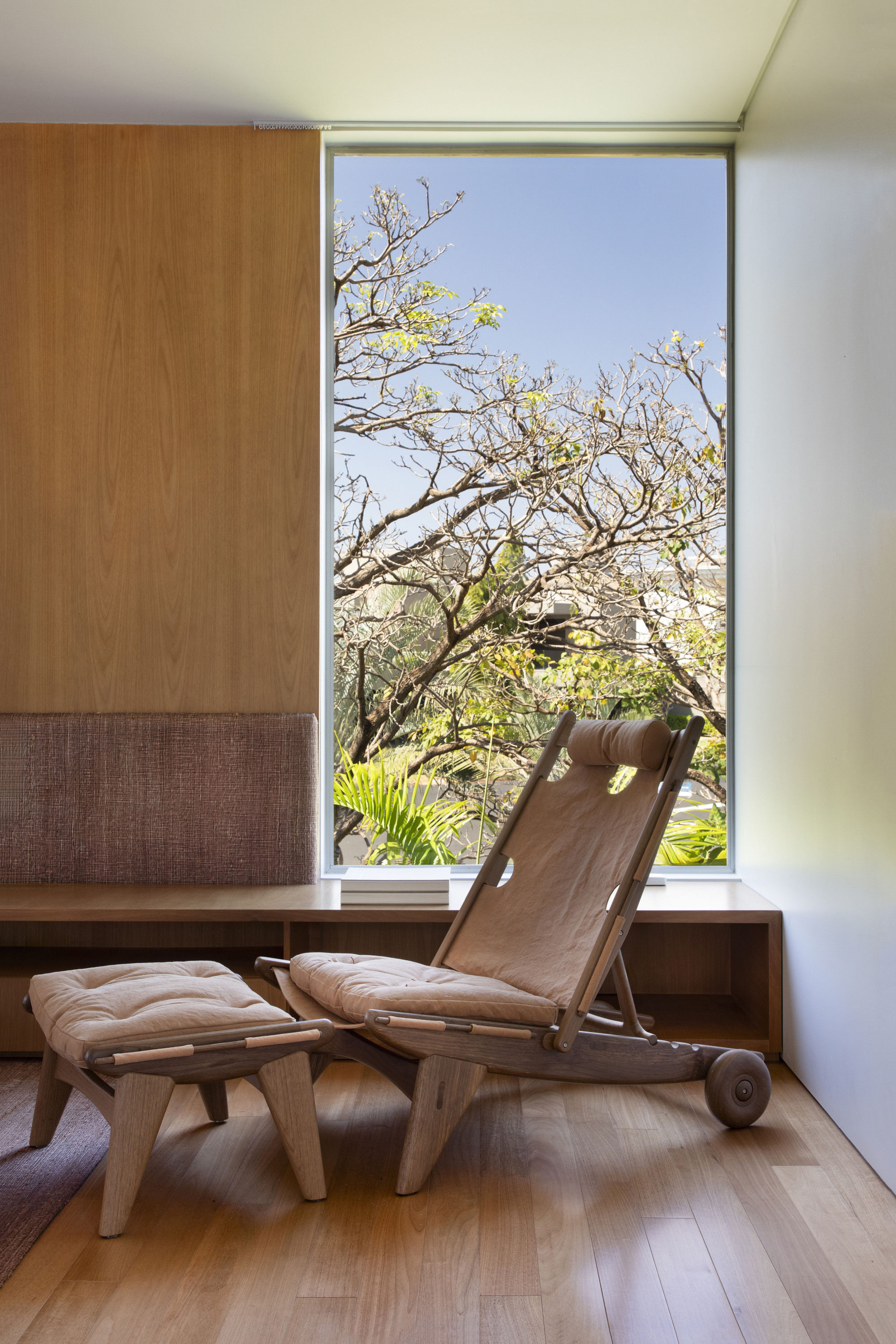 Wooden lounge chair and footstool beside a tall window framing a flowering tree against blue sky