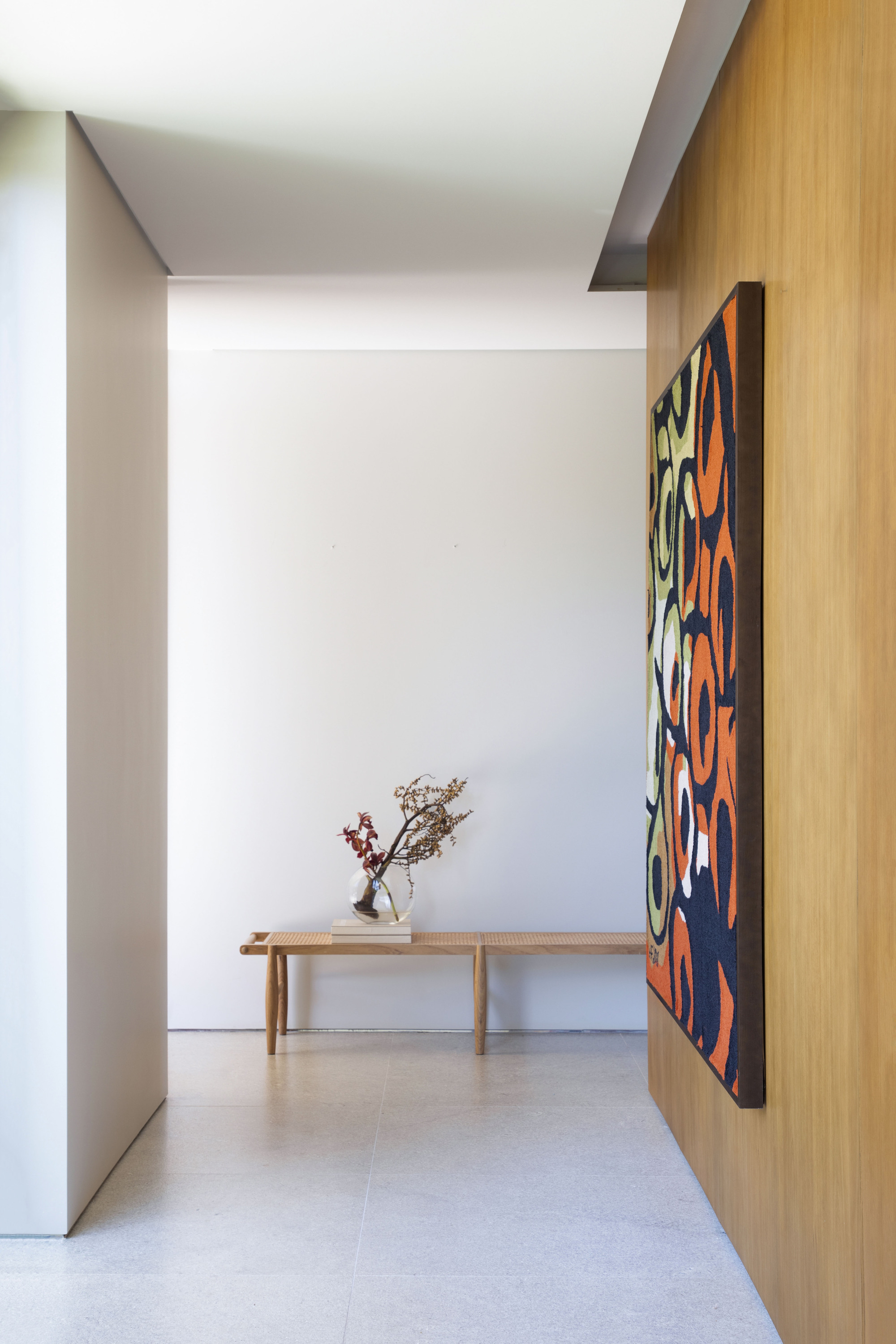 Entry corridor with a woven-seat wooden bench and dried branches in a glass vase beside a colorful abstract canvas