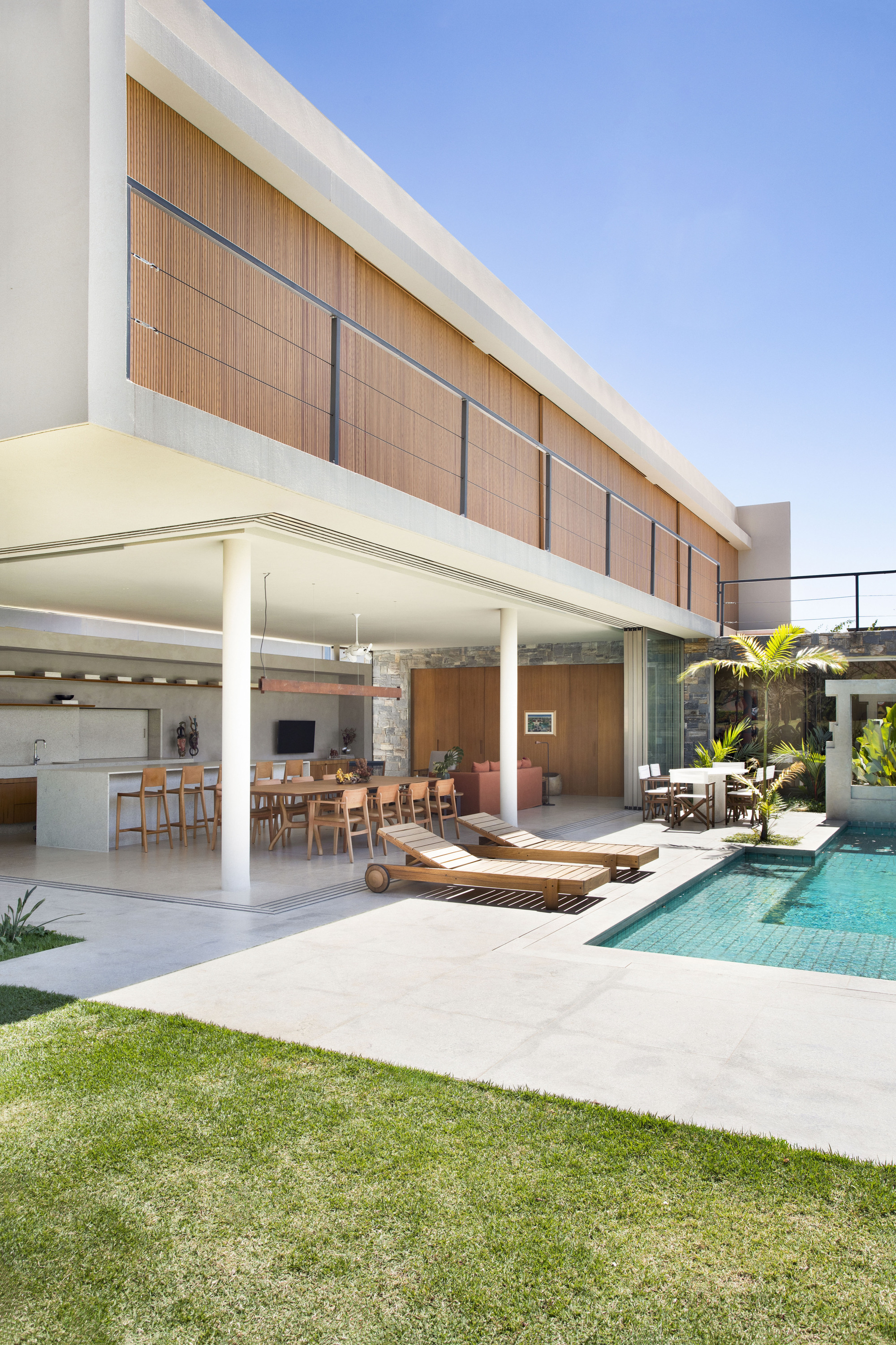 Rear facade with a timber-slatted upper volume above an open dining terrace, loungers and a tiled pool under clear blue sky