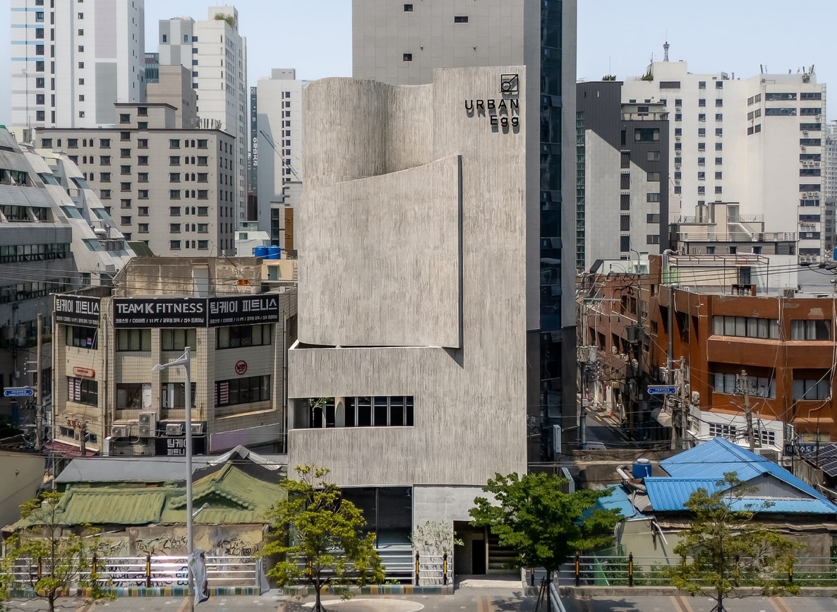 Frontal view of the curved board-marked concrete facade rising between low rooftops and apartment towers under a clear hazy sky