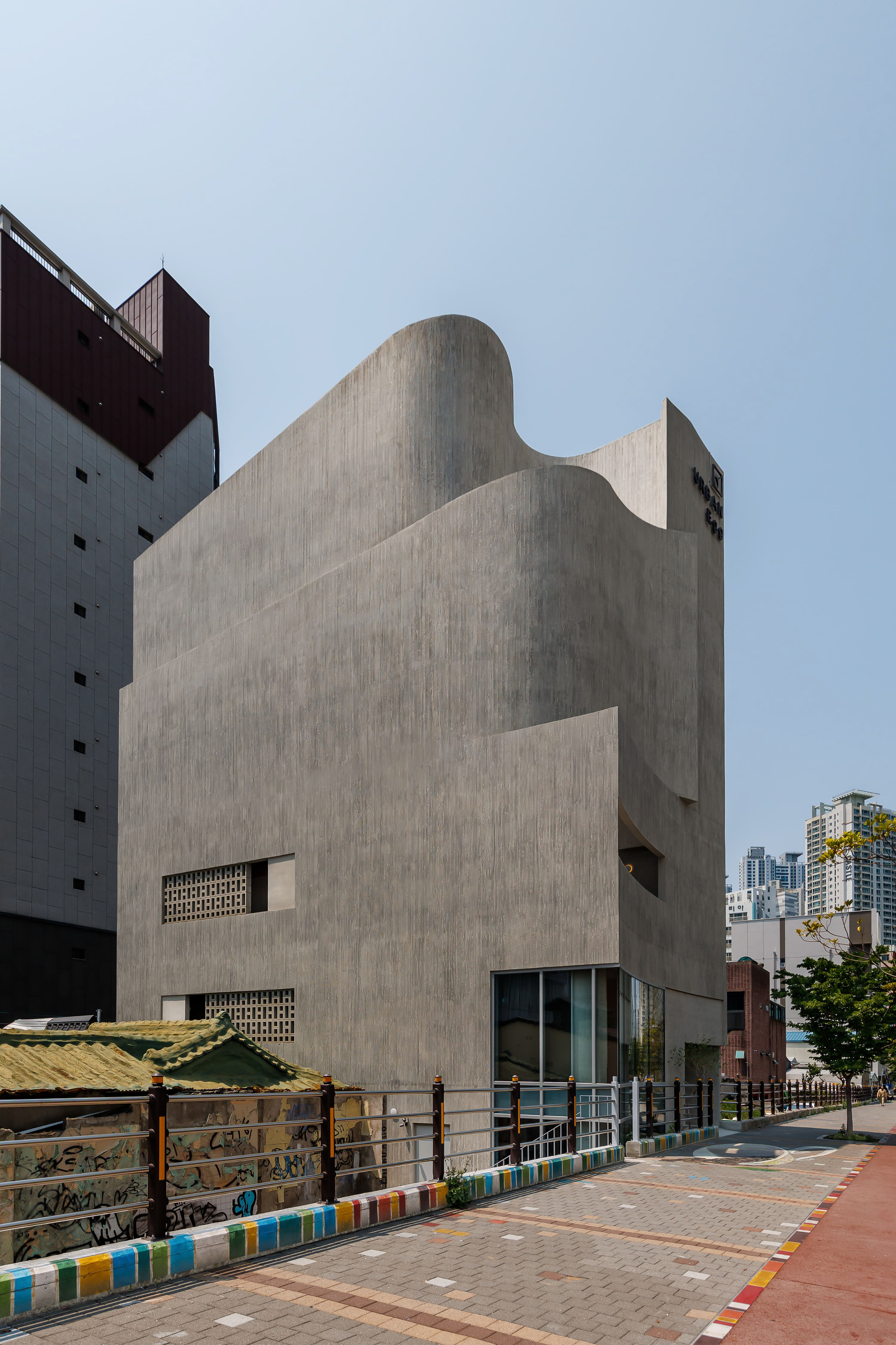 Corner view of the sculpted concrete volume with perforated block openings and a glazed ground floor beside a paved sidewalk