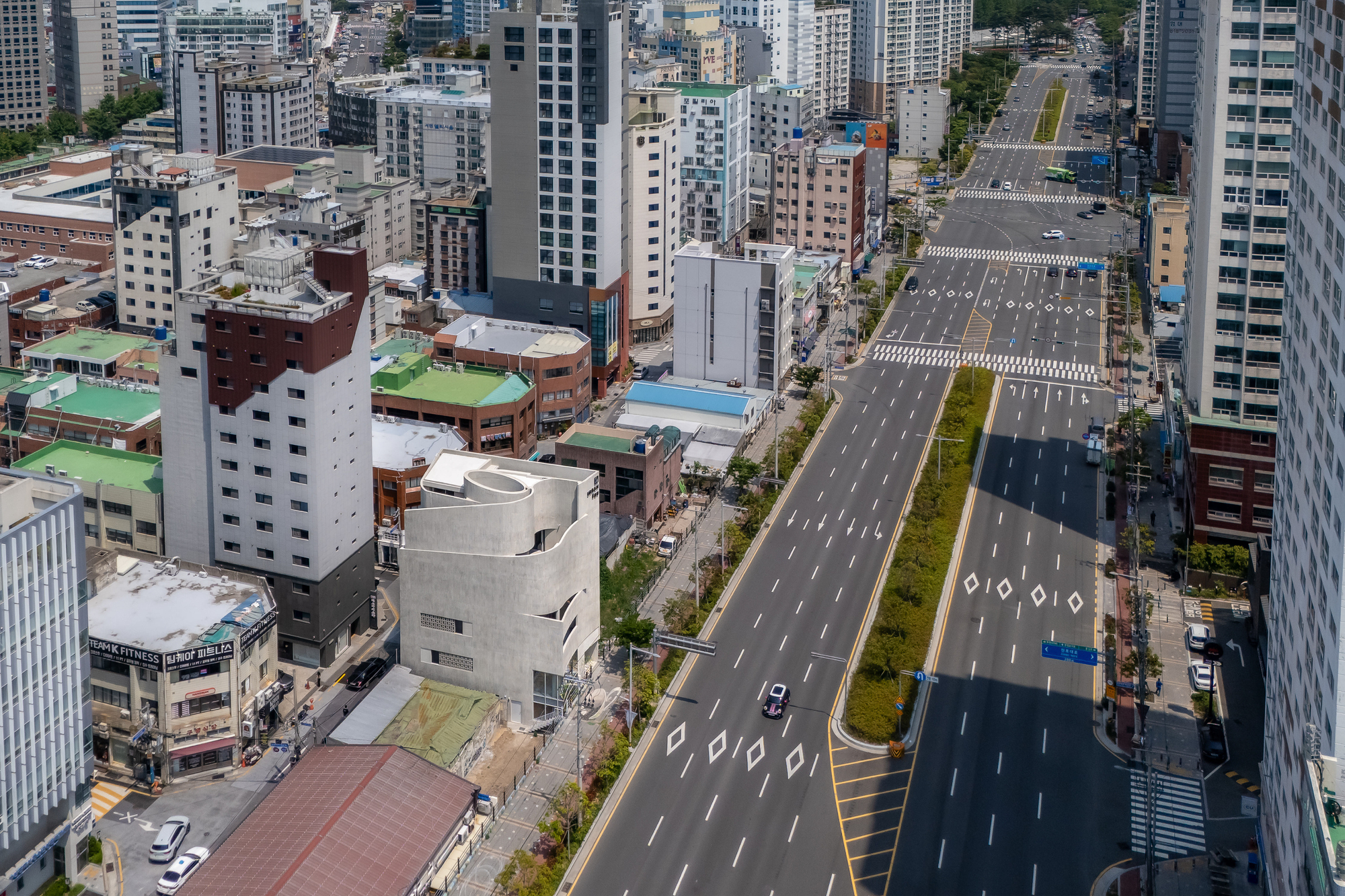 Elevated view of the pale curved building among mid-rise blocks beside a wide road with painted lane markings in daylight