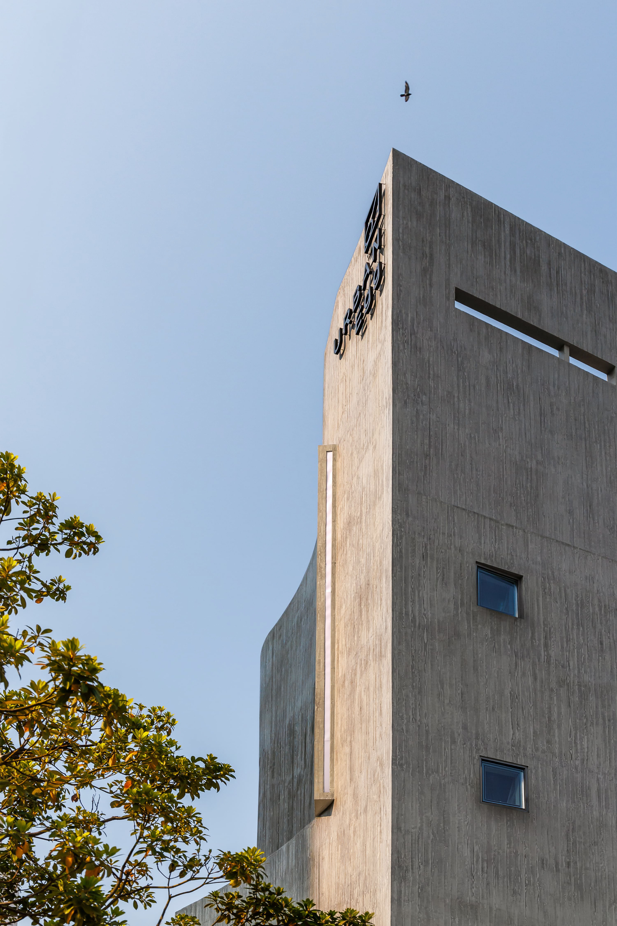 Board-formed concrete tower with a vertical light slot, small square windows and black lettering, a bird flying above and leaves in the foreground