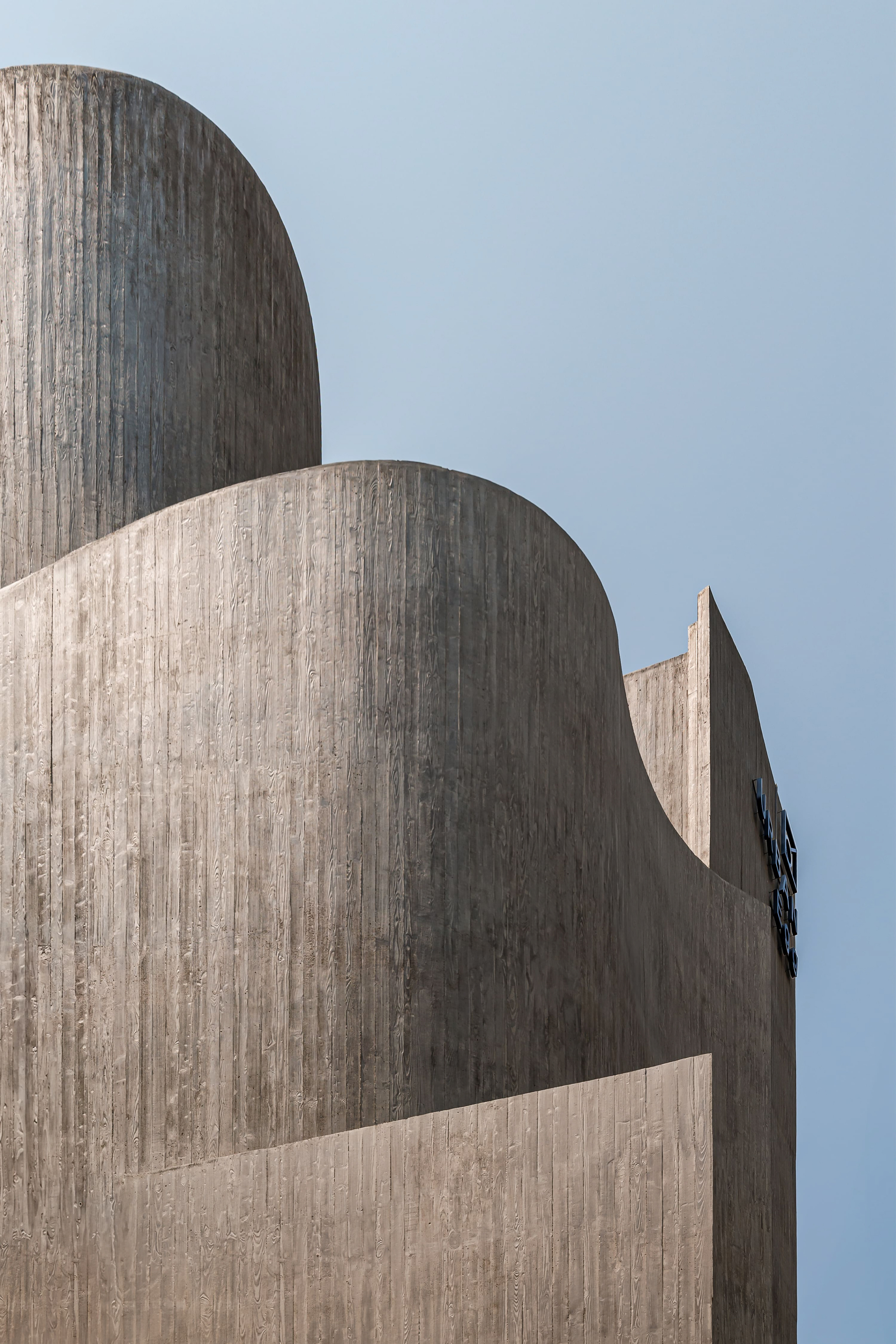 Close-up of stacked curved concrete walls showing vertical board-form texture against a pale blue sky