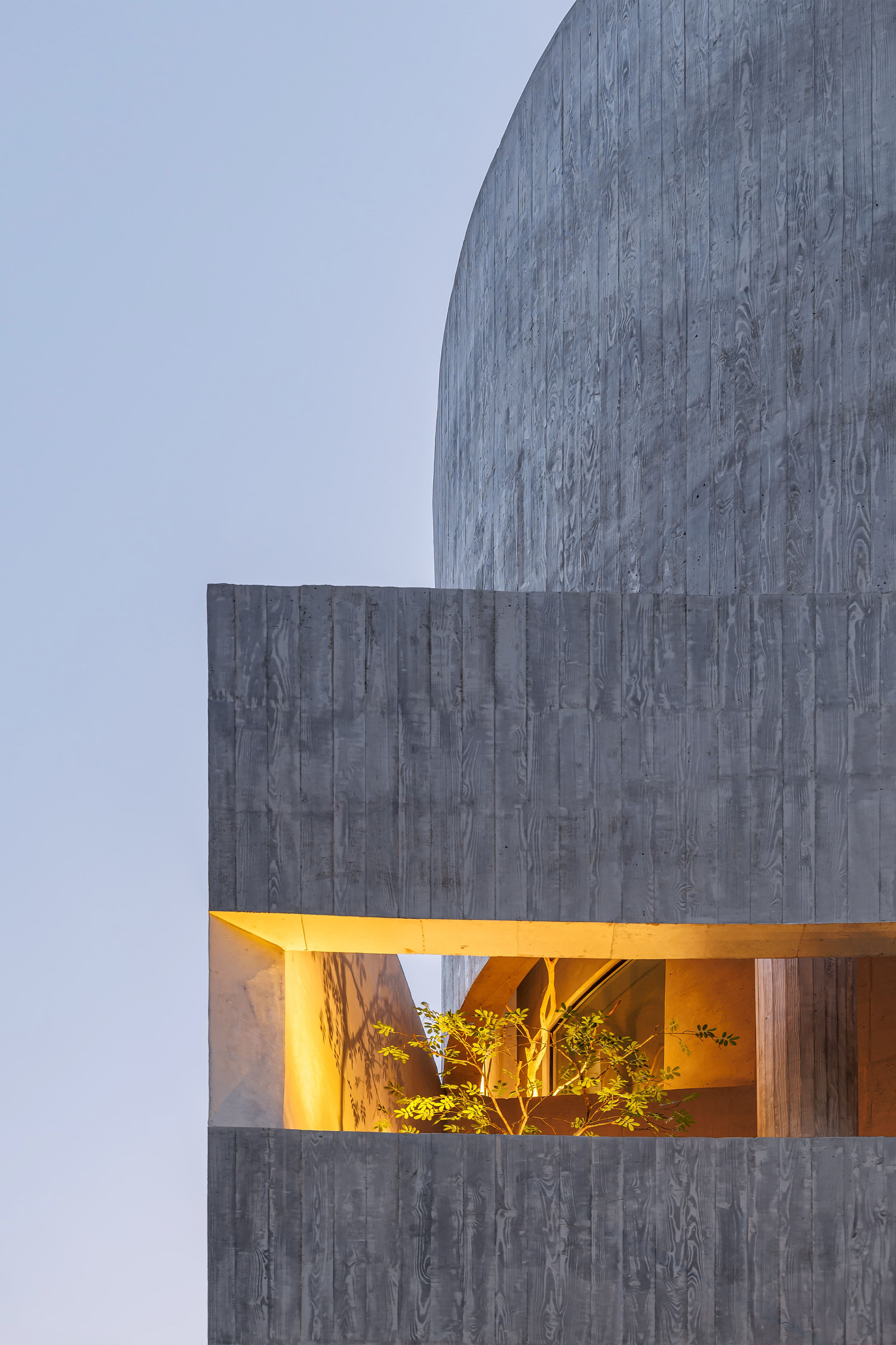 Corner of board-formed concrete volumes with a glowing slot window and a small leafy tree at dusk