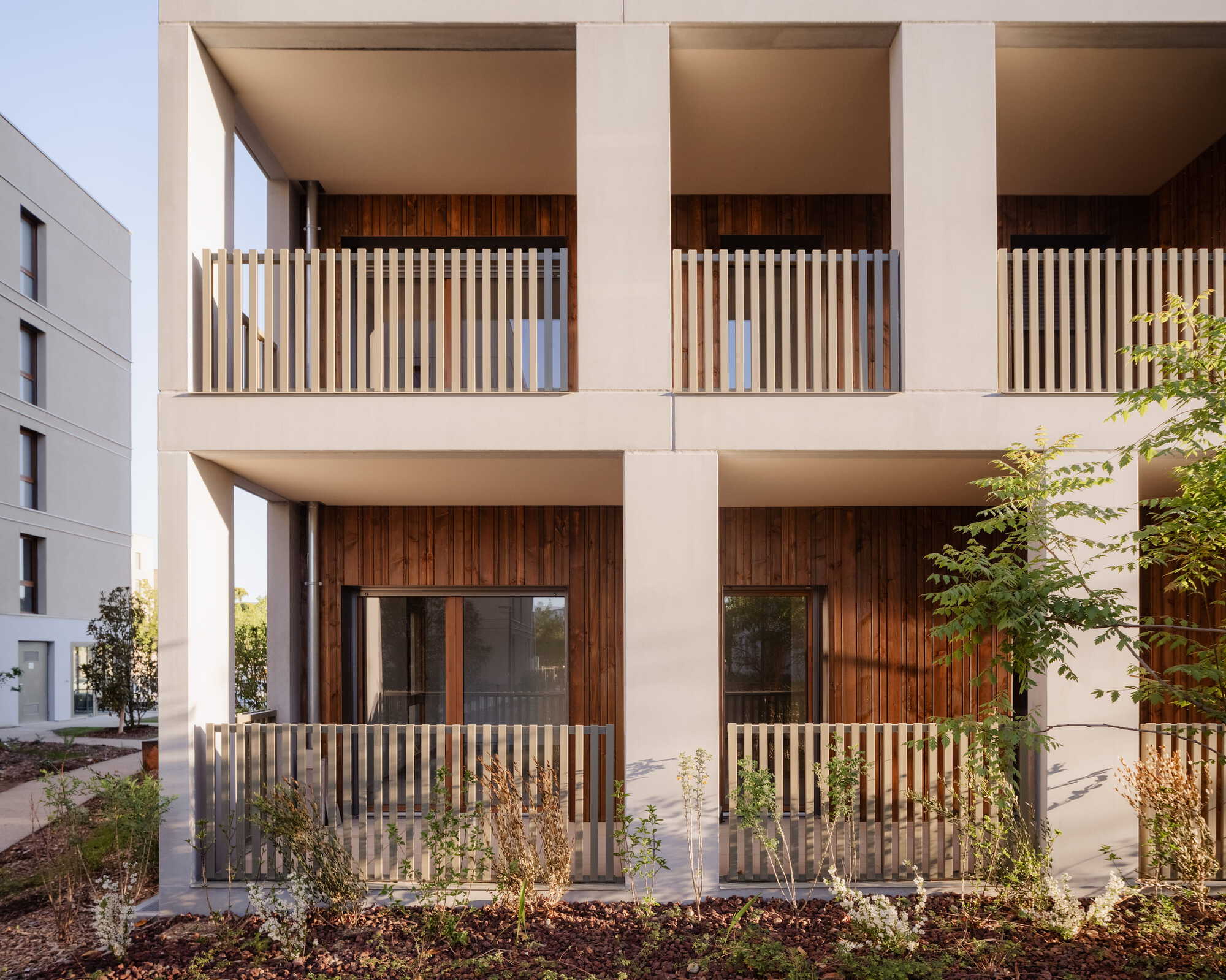 Ground and first floor loggias with square concrete piers, slatted balustrades and vertical timber cladding above a mulched planting bed
