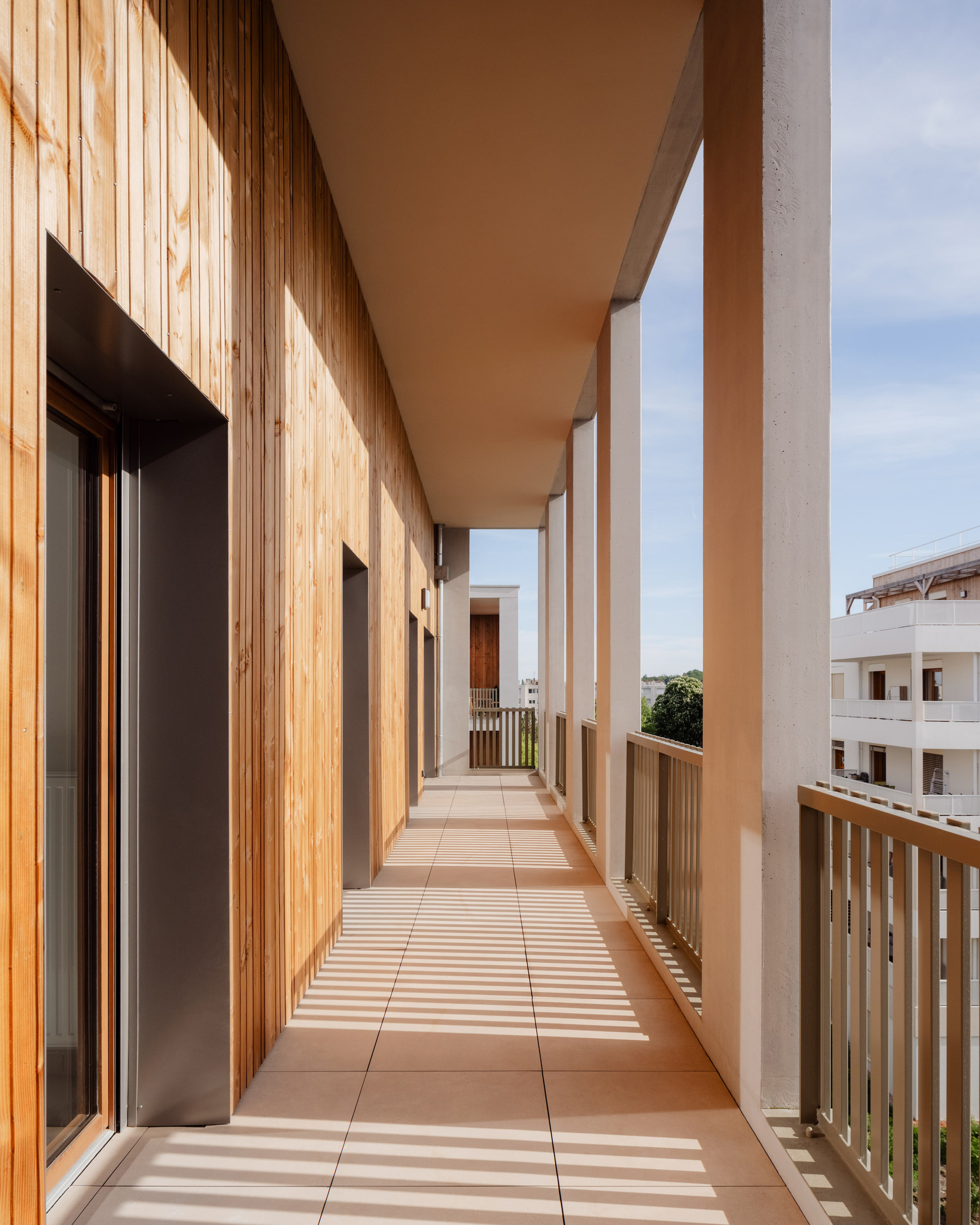 Covered balcony walkway with vertical timber cladding, slatted railings and sun stripes falling across the terracotta floor tiles