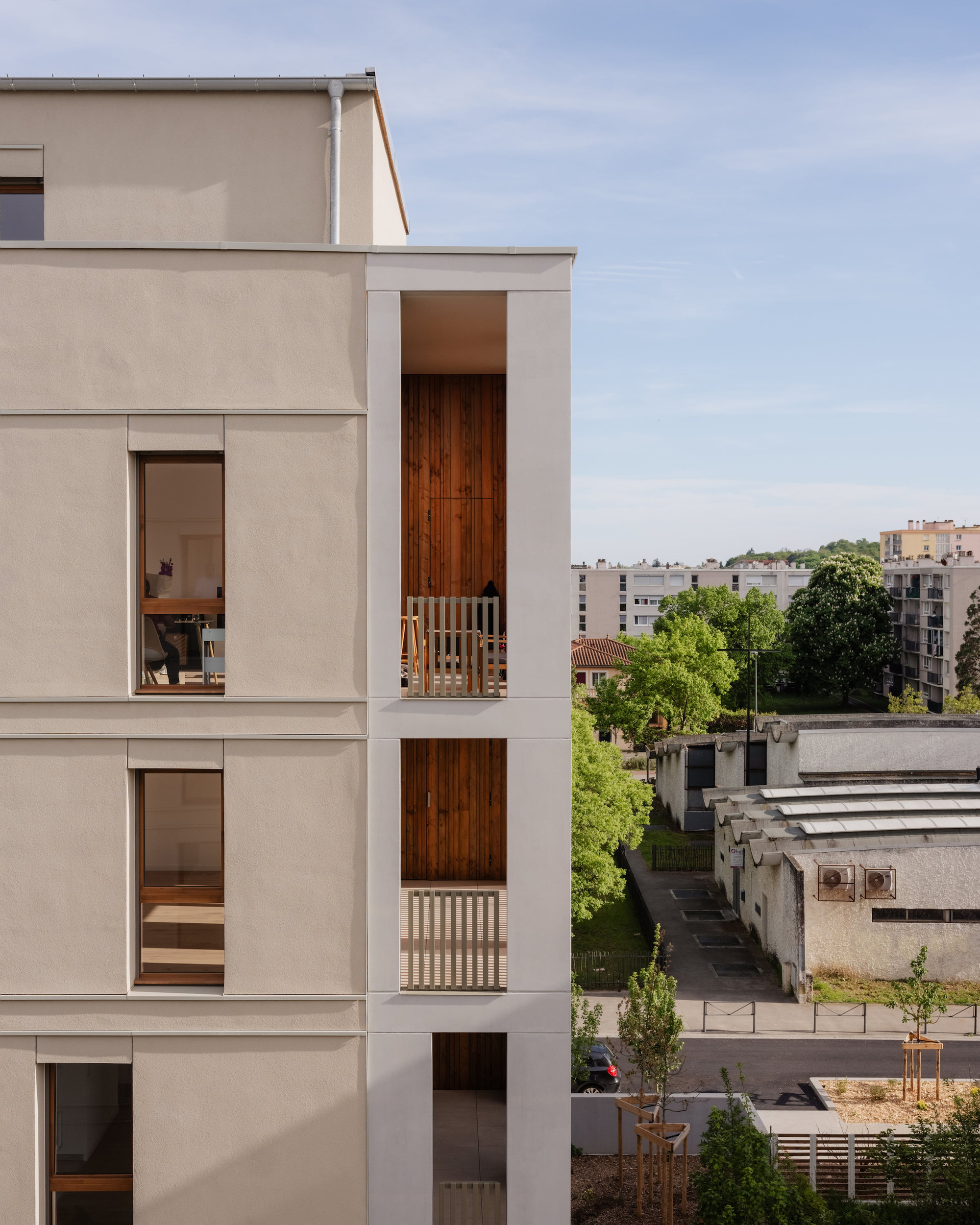 Side elevation of a rendered apartment block with stacked timber-lined loggias, overlooking low industrial sheds and street trees under a pale sky