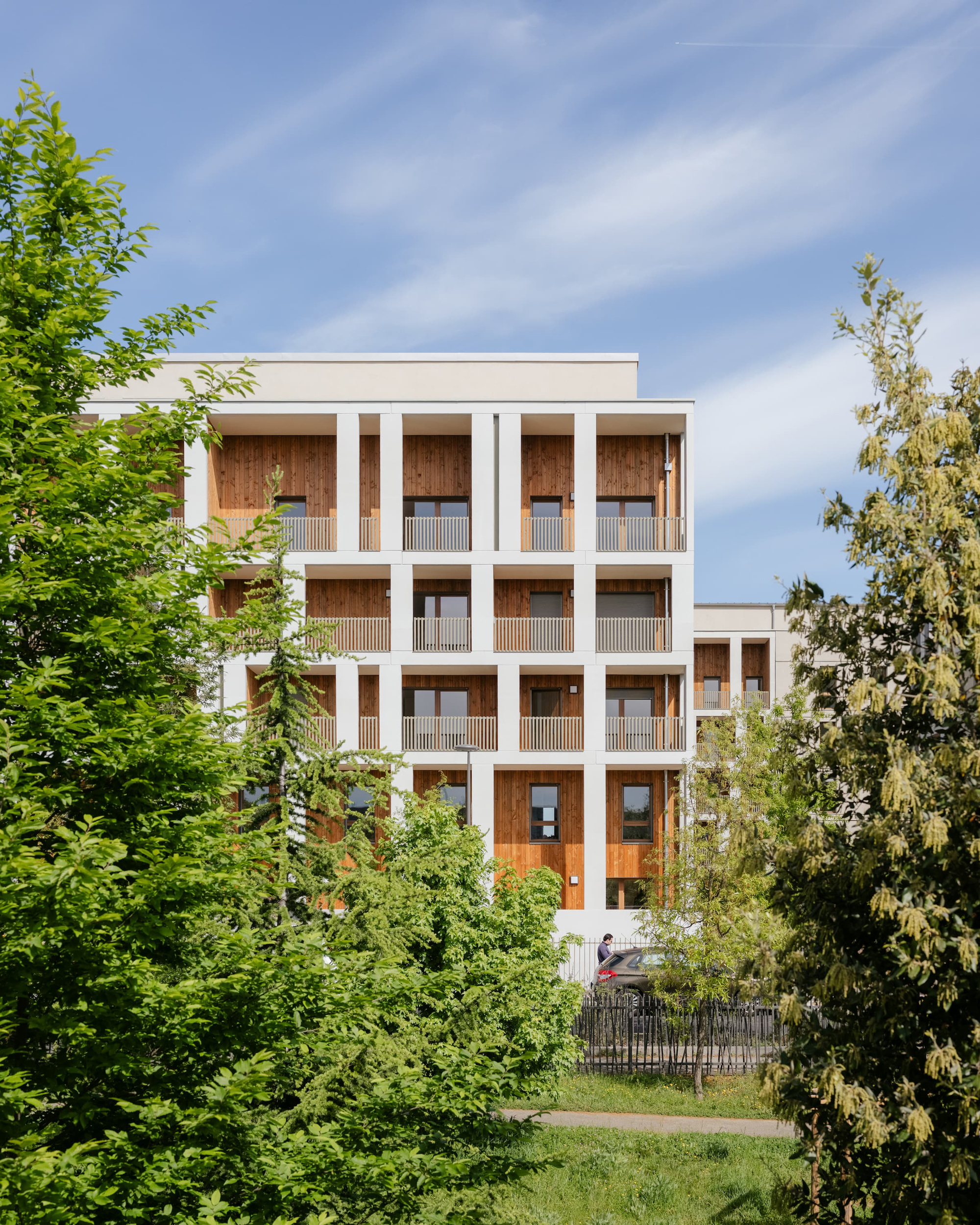 Facade of white concrete columns framing timber-lined balconies with slatted railings, seen between trees under wispy clouds