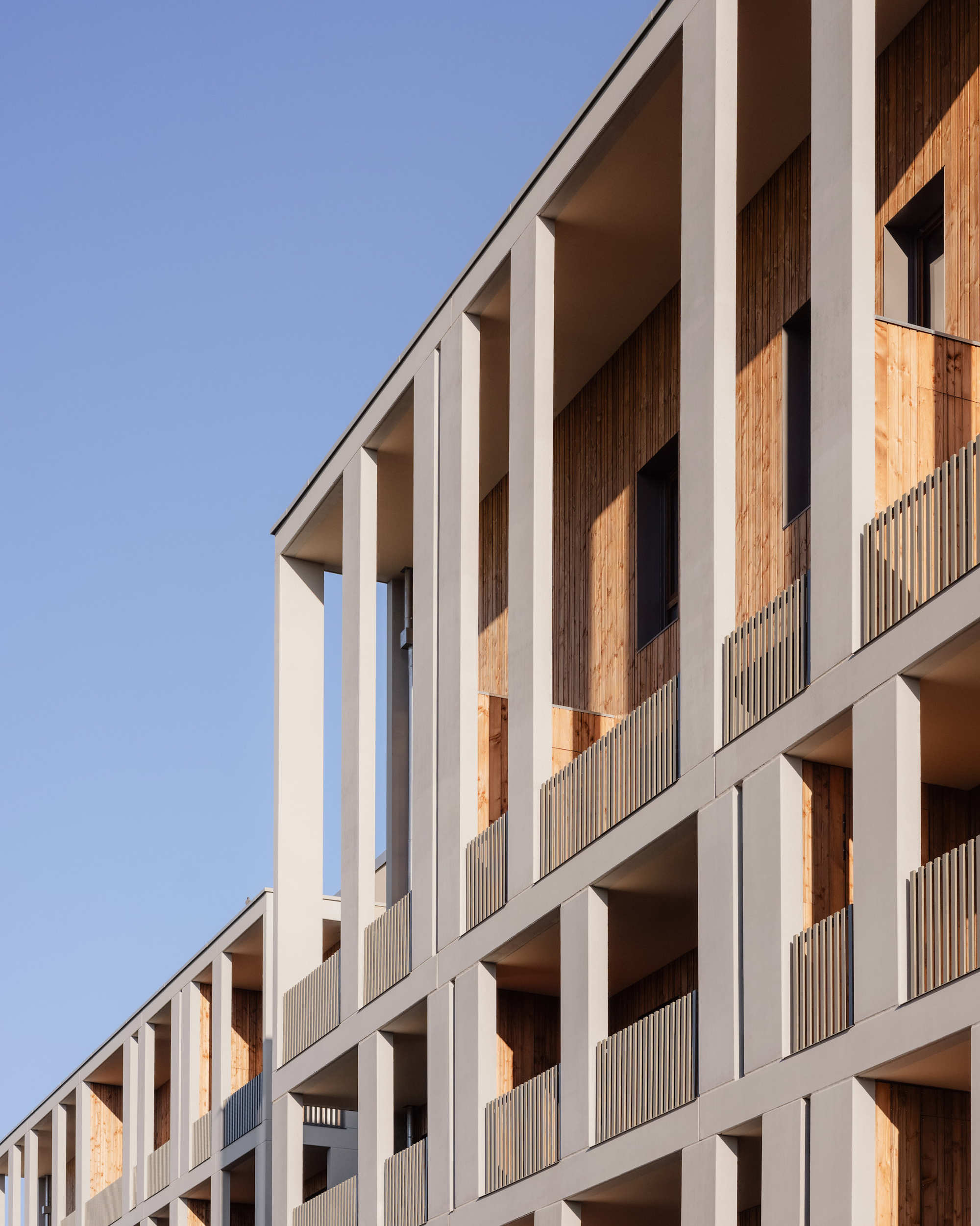 Upward view of the concrete grid facade with deep loggias, vertical timber boards and slatted balustrades against a clear blue sky