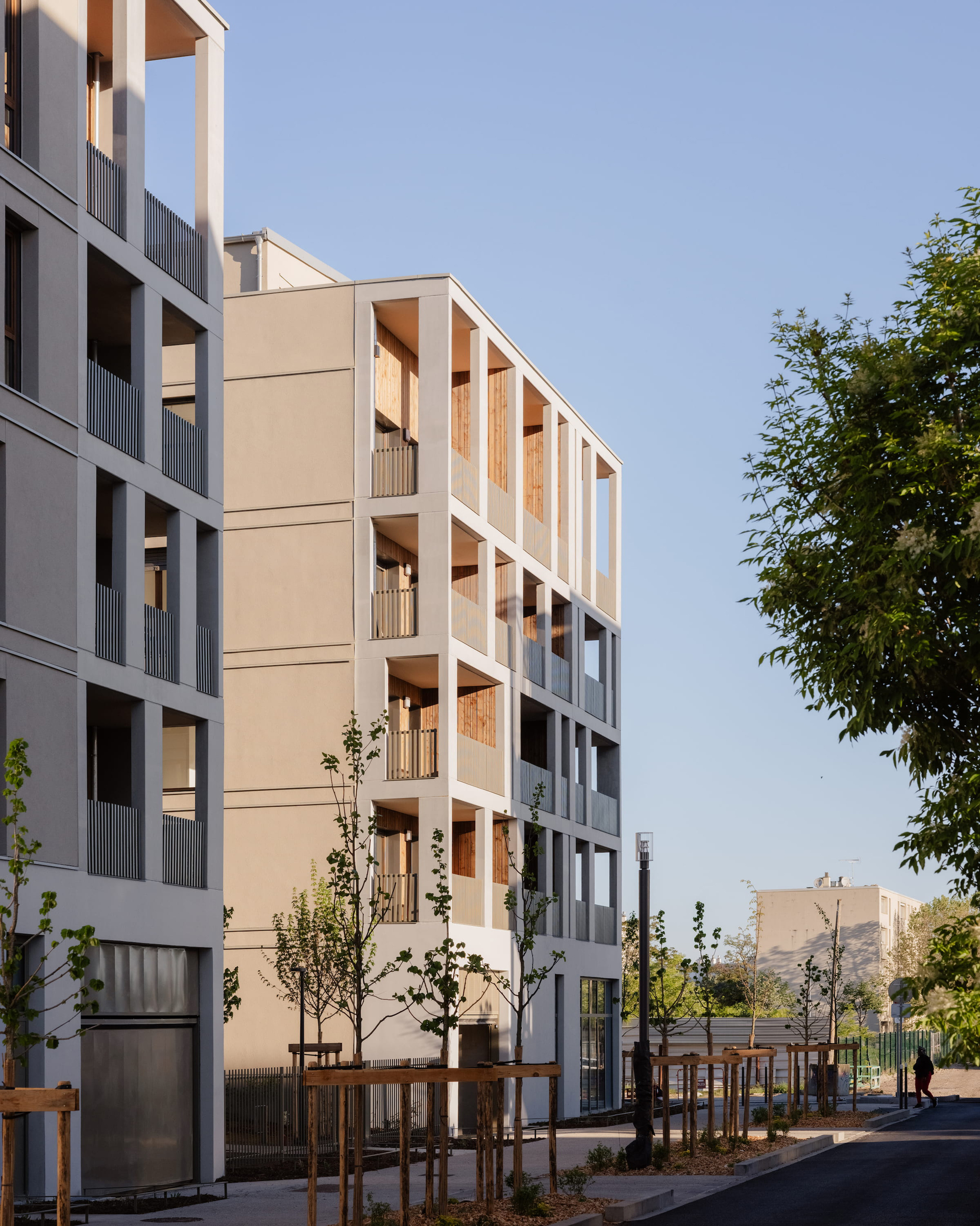 Street view of two stepped apartment blocks with open timber loggias and young saplings in wooden supports at dusk