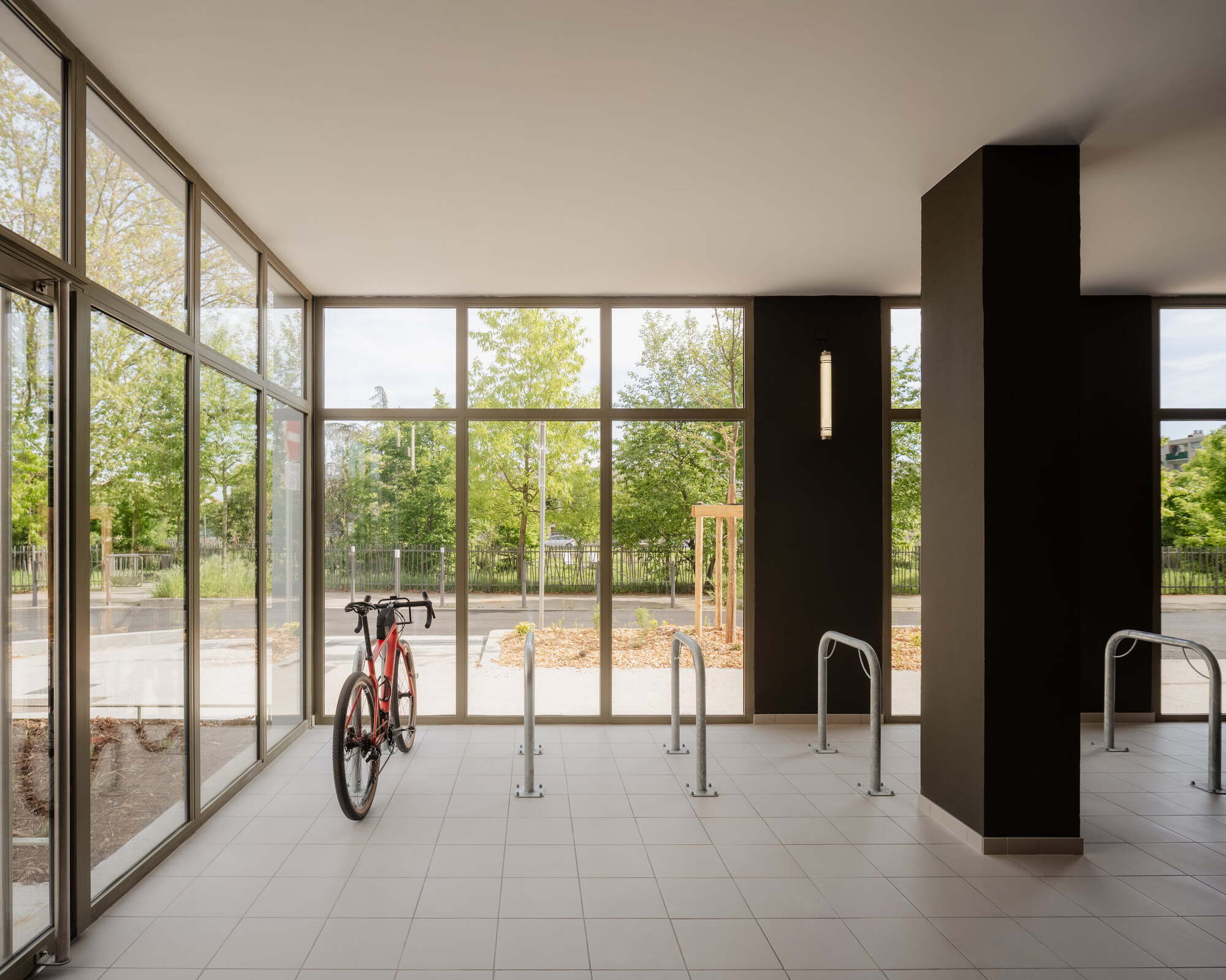 Ground-floor bike room with glazed walls, steel racks and a dark column, one red bicycle parked and trees outside
