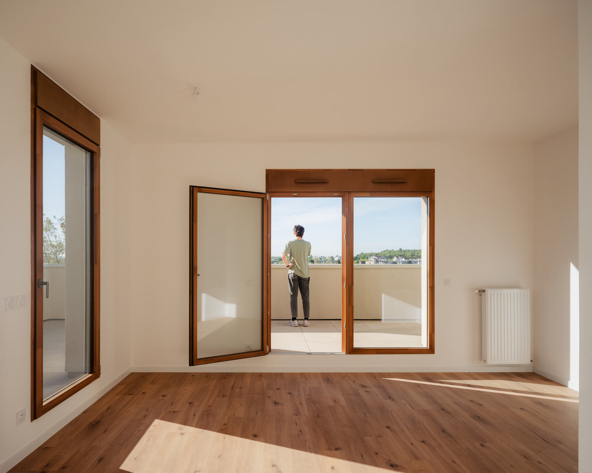 Empty room with timber-framed glazed doors open to a terrace, where a person looks out over distant treetops in afternoon sun