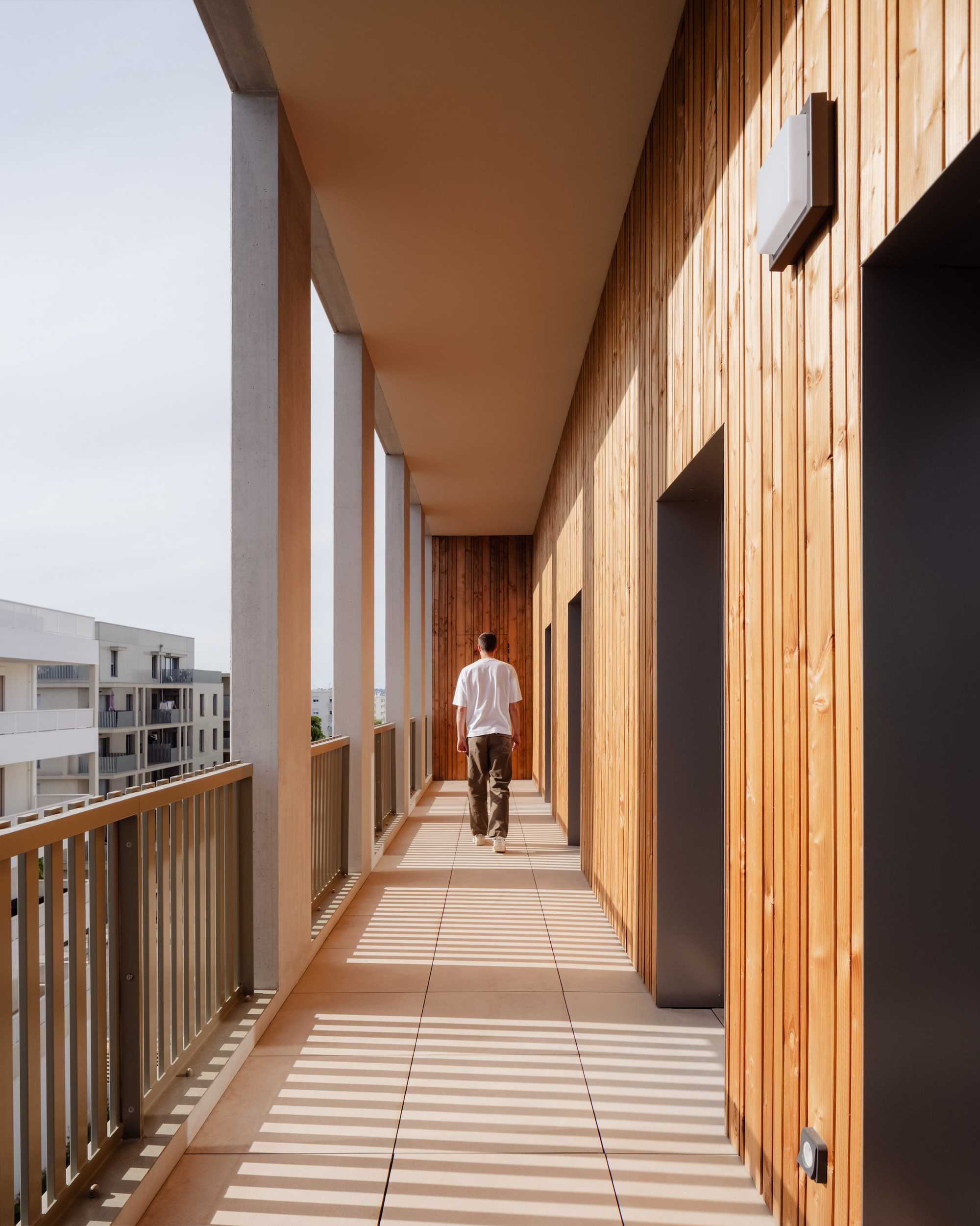 Person walking along a covered balcony walkway beside vertical timber cladding, with slatted shadows falling across pale paving tiles
