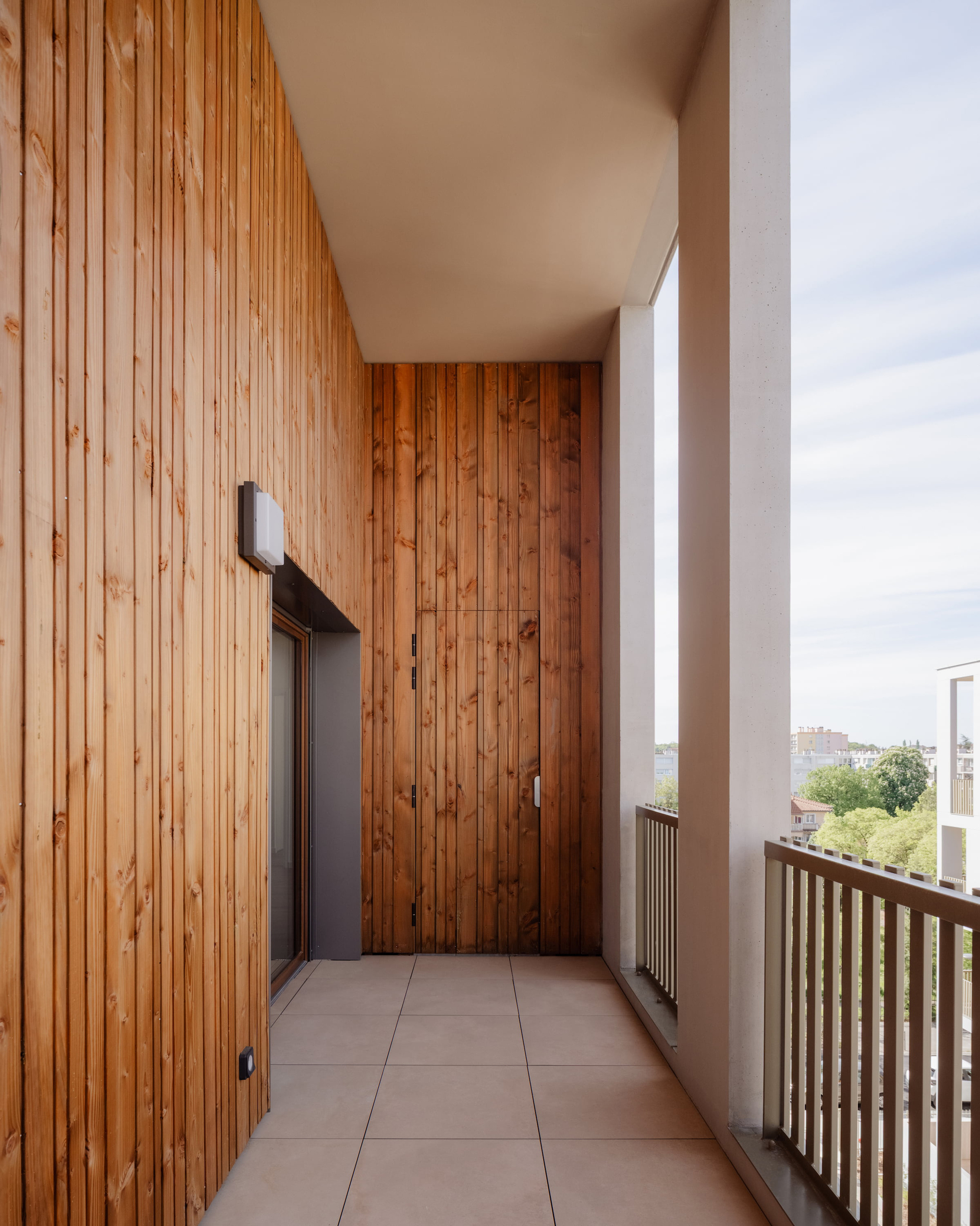 Recessed balcony corner with vertical larch cladding, a flush storage door and a slim railing overlooking trees under soft cloud