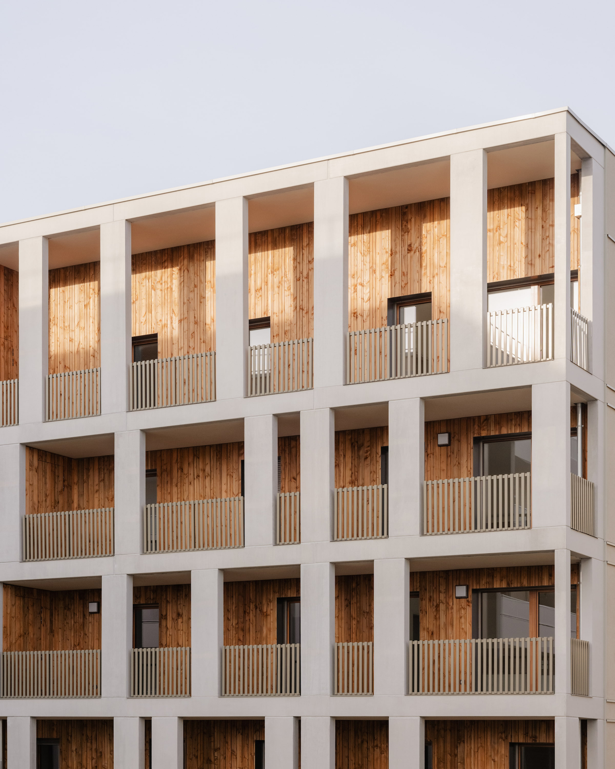 Facade grid of pale concrete frames holding timber-clad loggias with slatted balustrades against an overcast sky