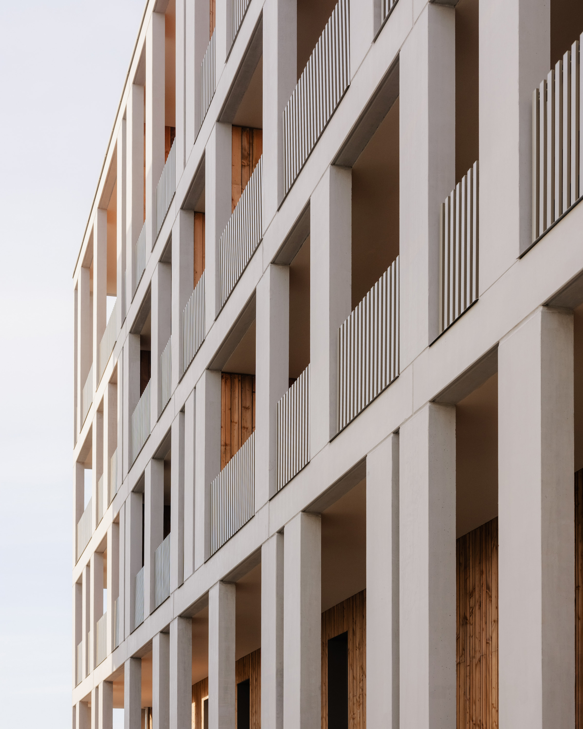 Angled close view of concrete columns and slatted balcony balustrades receding up the facade, with timber glimpsed inside the loggias in pale light