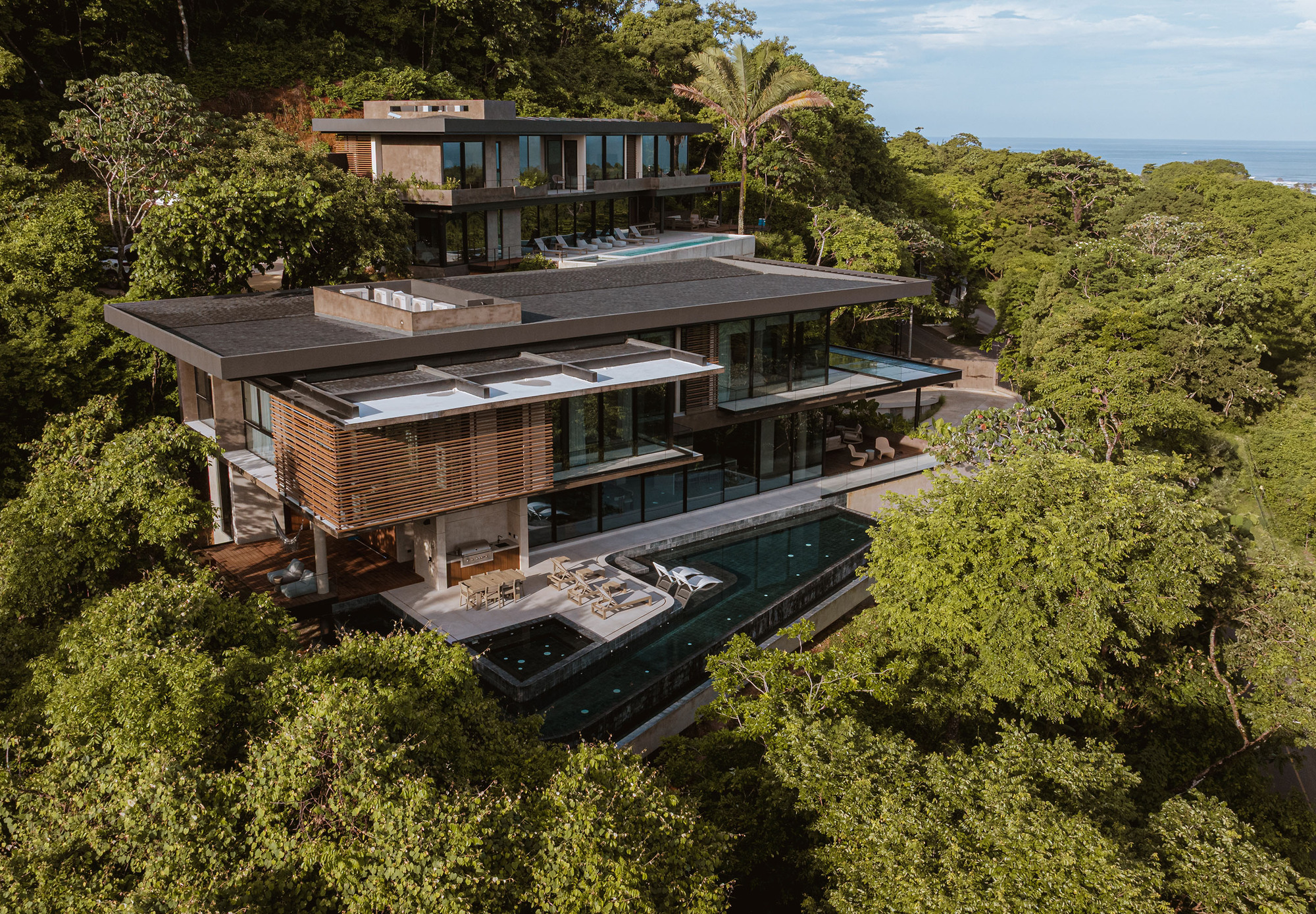 Aerial view of stacked flat-roofed volumes with timber louvers and an infinity pool cutting through dense green forest canopy