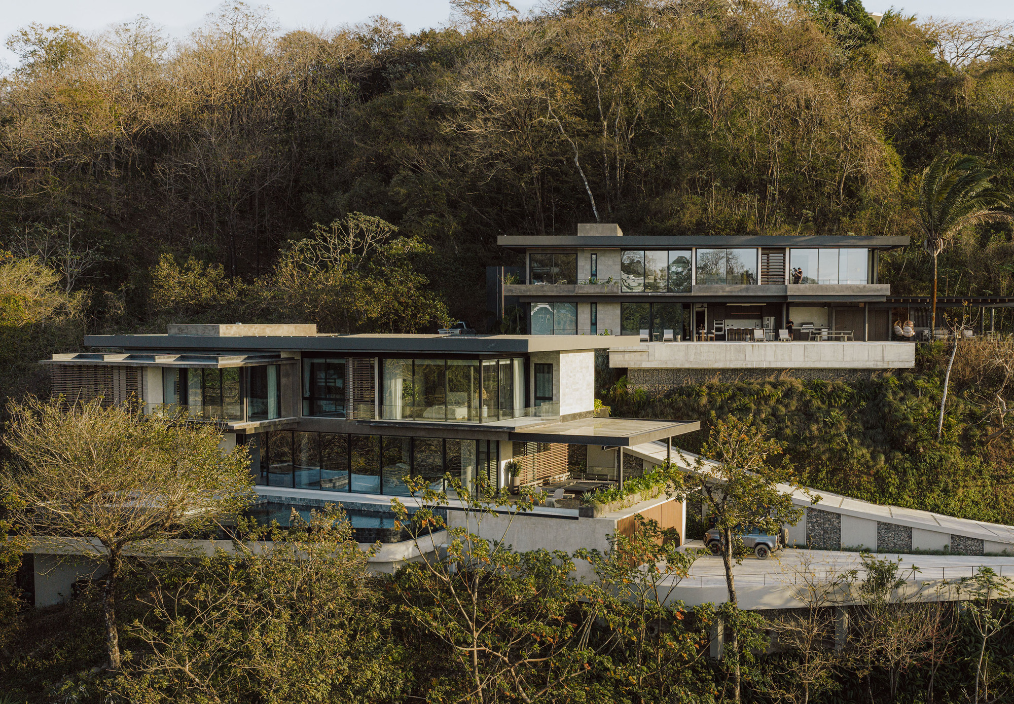 Terraced glass and concrete volumes on a forested hillside in warm late afternoon light, with a ramp curving below