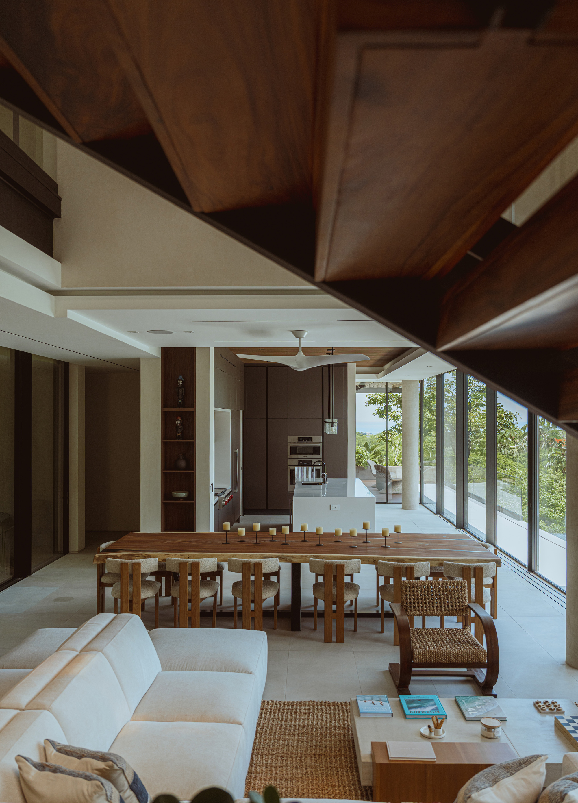 Open living and dining space seen beneath a dark timber stair, with a long wood table and floor-to-ceiling glazing