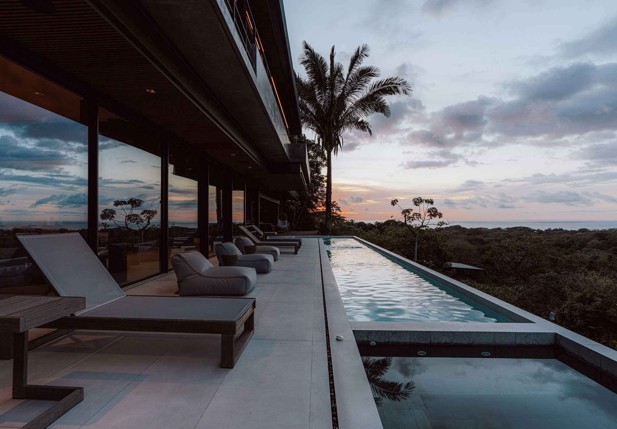 Pool terrace with lounge chairs along a glazed wall, a palm tree and a dusk sky over the distant sea