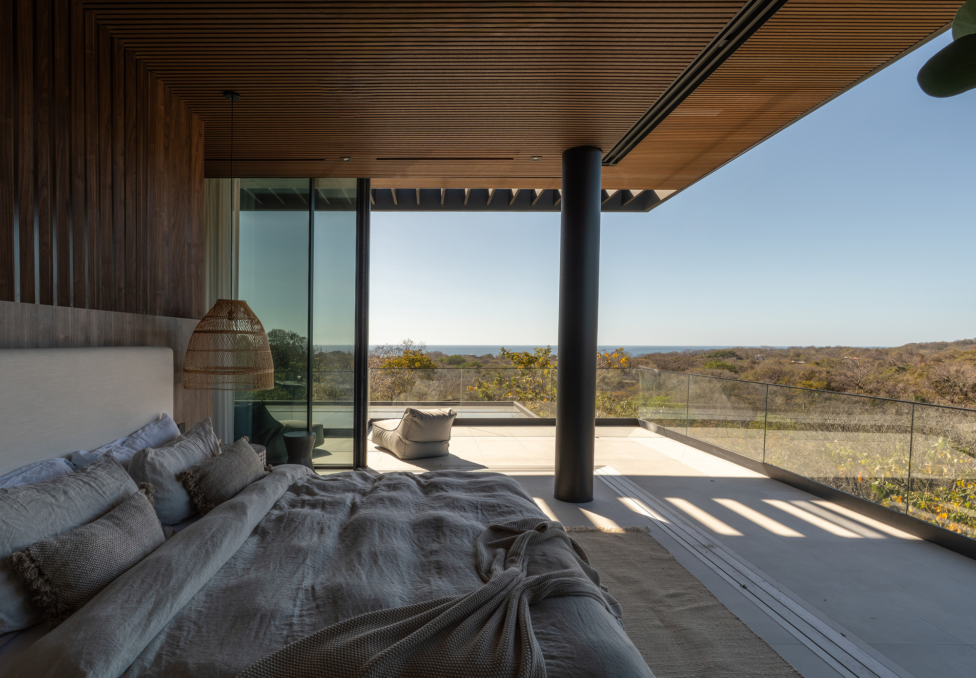 Bedroom opening onto a terrace with a black steel column, slatted timber ceiling and glass balustrade in bright sun