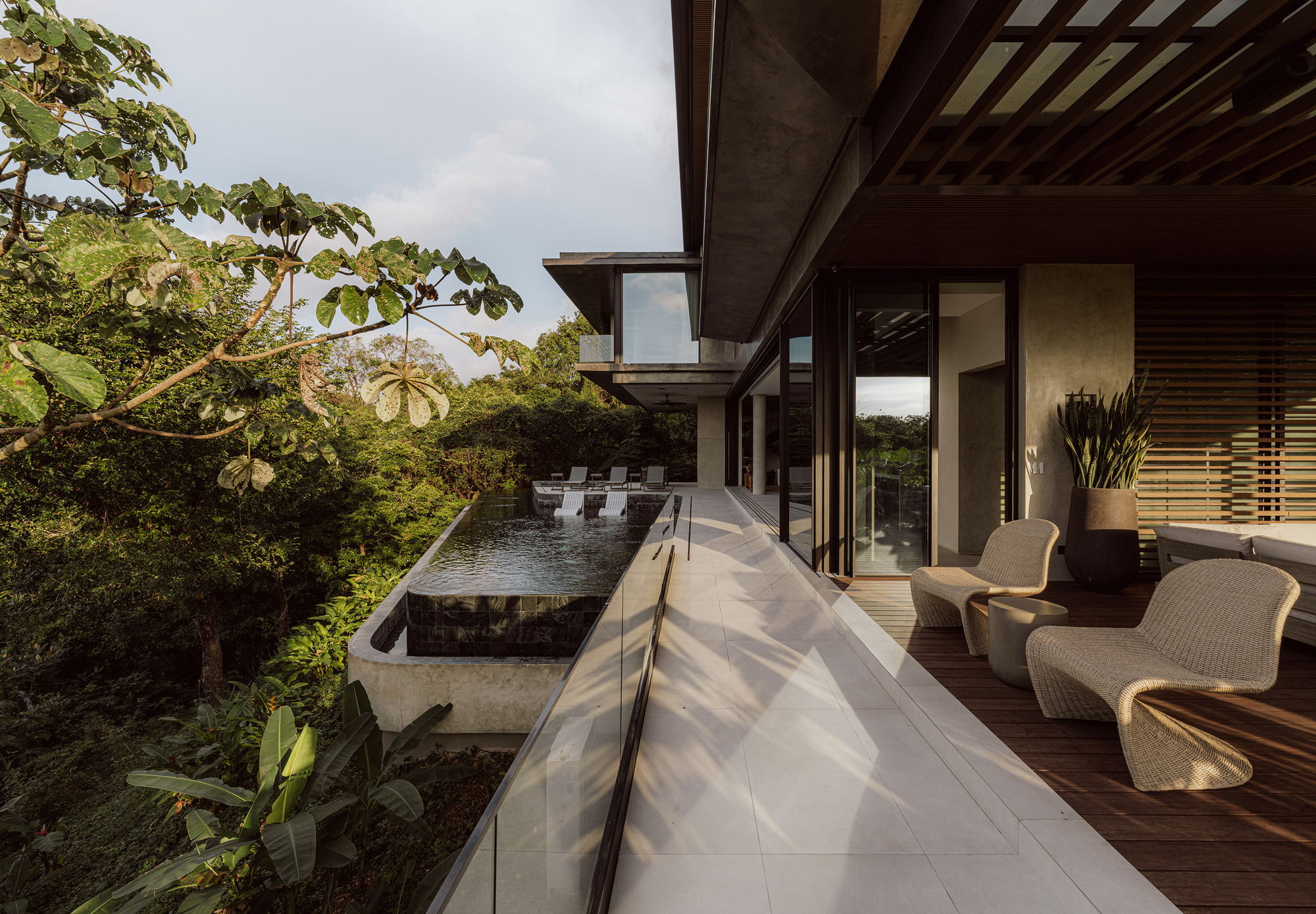 Stone-paved walkway beside a glass balustrade, with wicker chairs on a timber deck and a pool among the trees