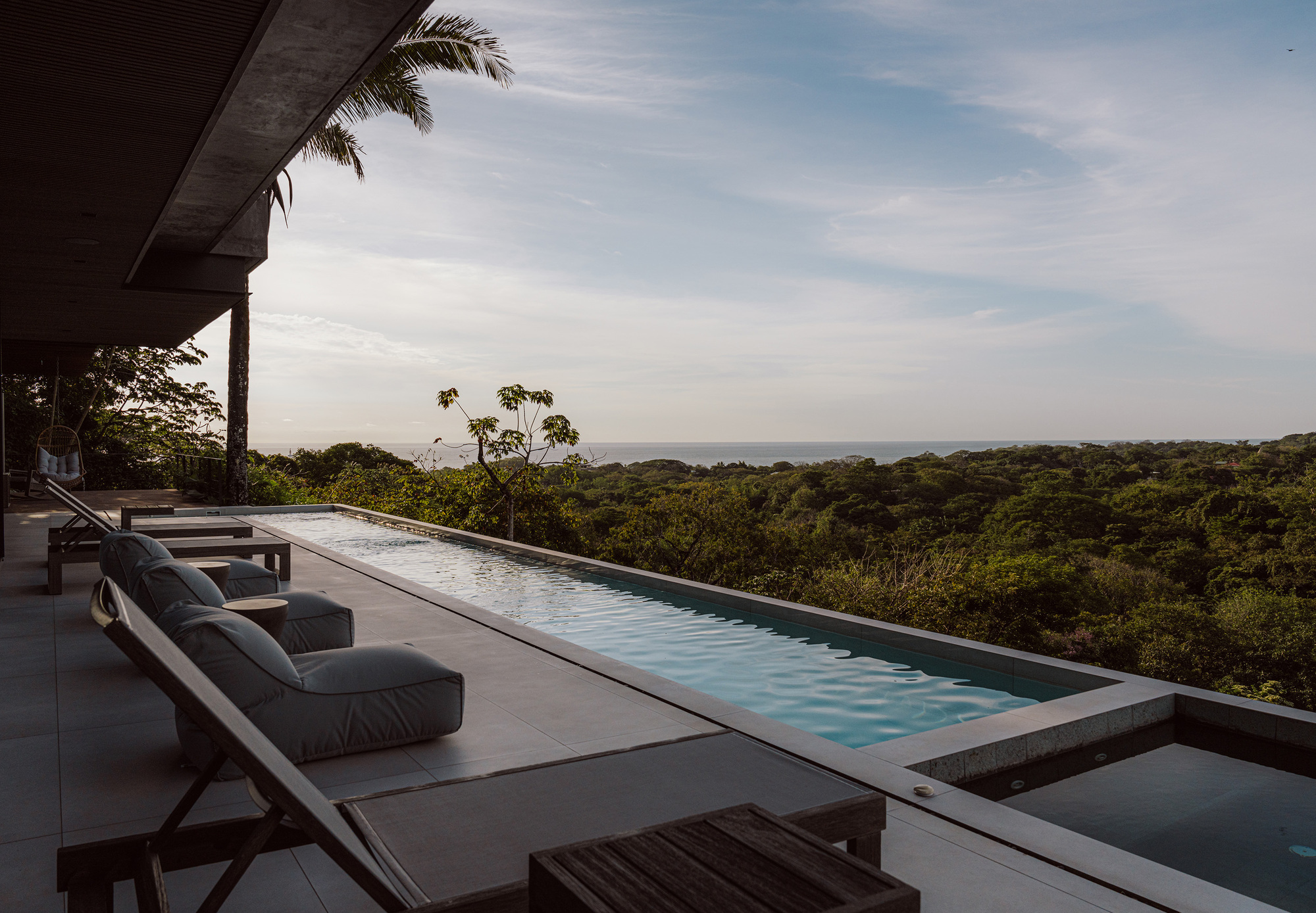 Long infinity pool with lounge chairs on a terrace overlooking forest canopy and distant ocean under soft evening clouds