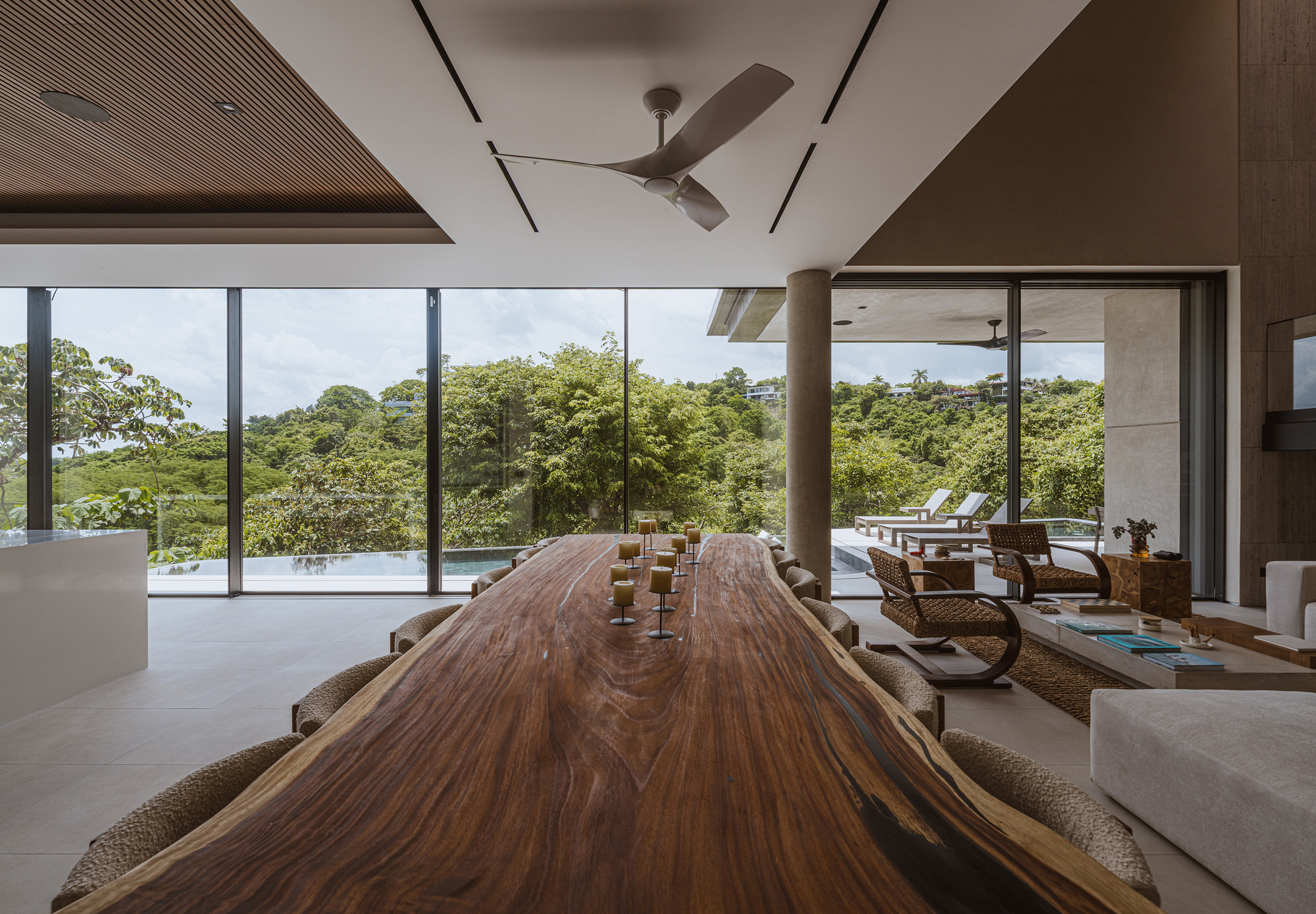 Long live-edge wood dining table facing floor-to-ceiling glass that opens onto a pool and green hillside