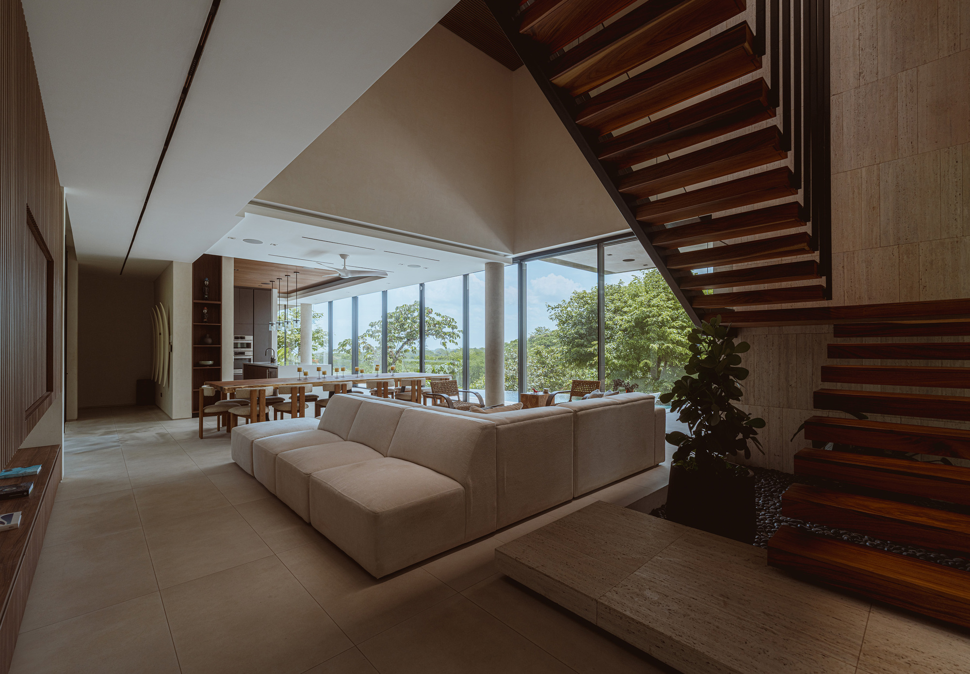 Living room with a pale sectional sofa beside a floating timber staircase, glass wall and treetops beyond