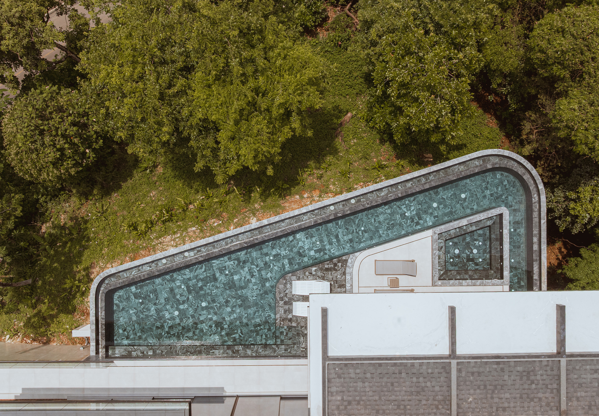 Top-down view of an angular mosaic-tiled pool edged in stone, set against a steep wooded slope