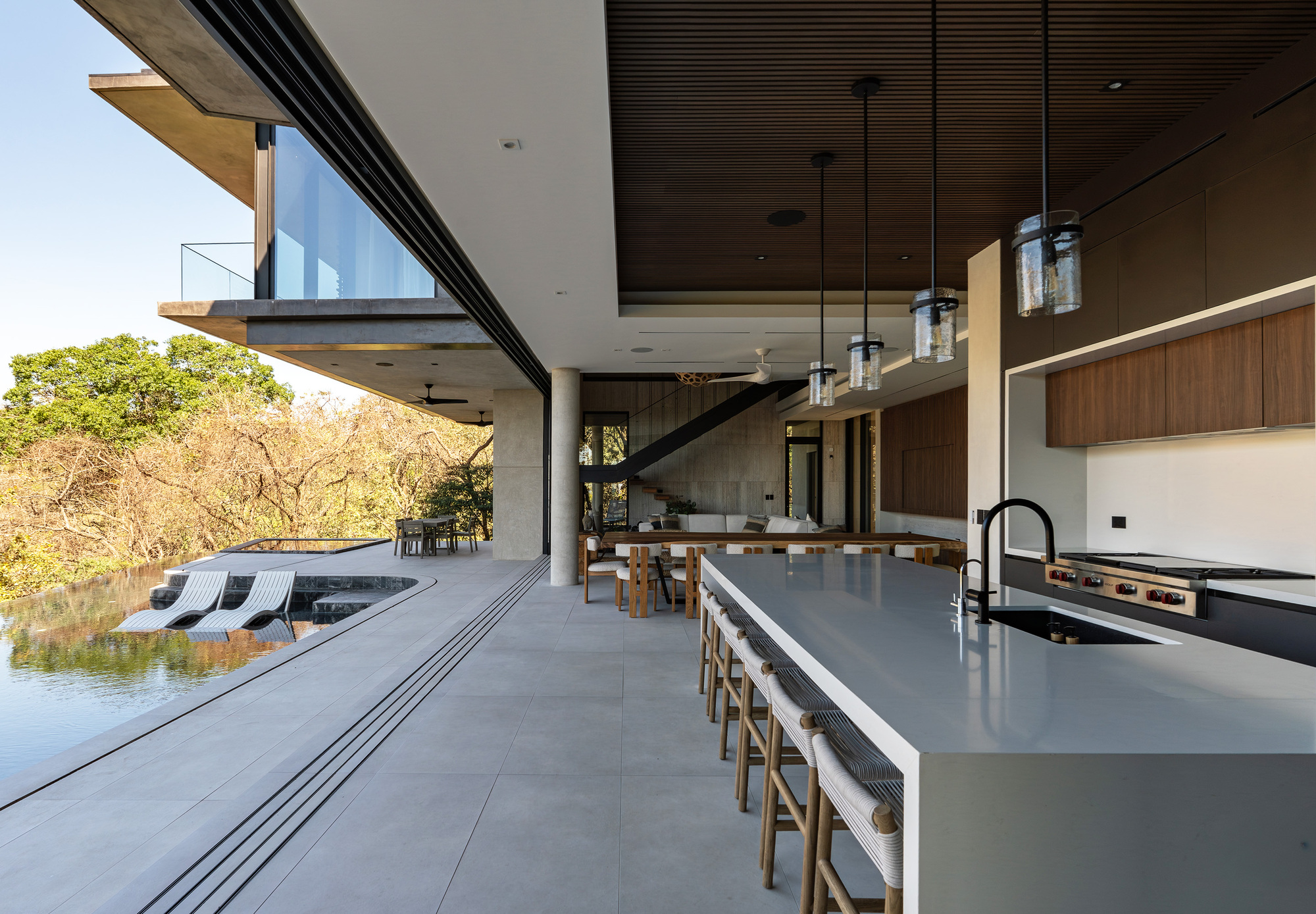 Open kitchen island with bar stools under a slatted timber ceiling, sliding glass leading to a pool terrace in daylight