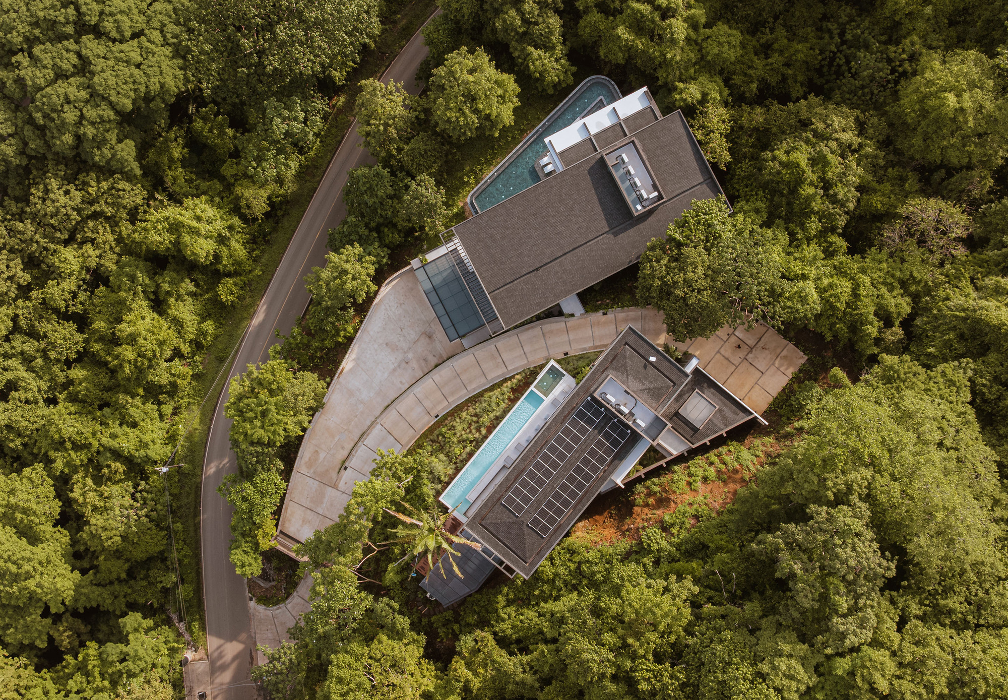 Drone view of two pitched-roof volumes with pools and solar panels, linked by a curving drive through dense green forest