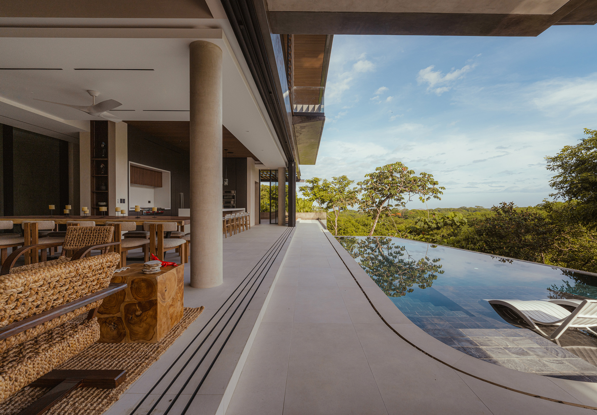 Covered terrace with a round-edged infinity pool and woven lounge chairs facing treetops under a partly cloudy sky