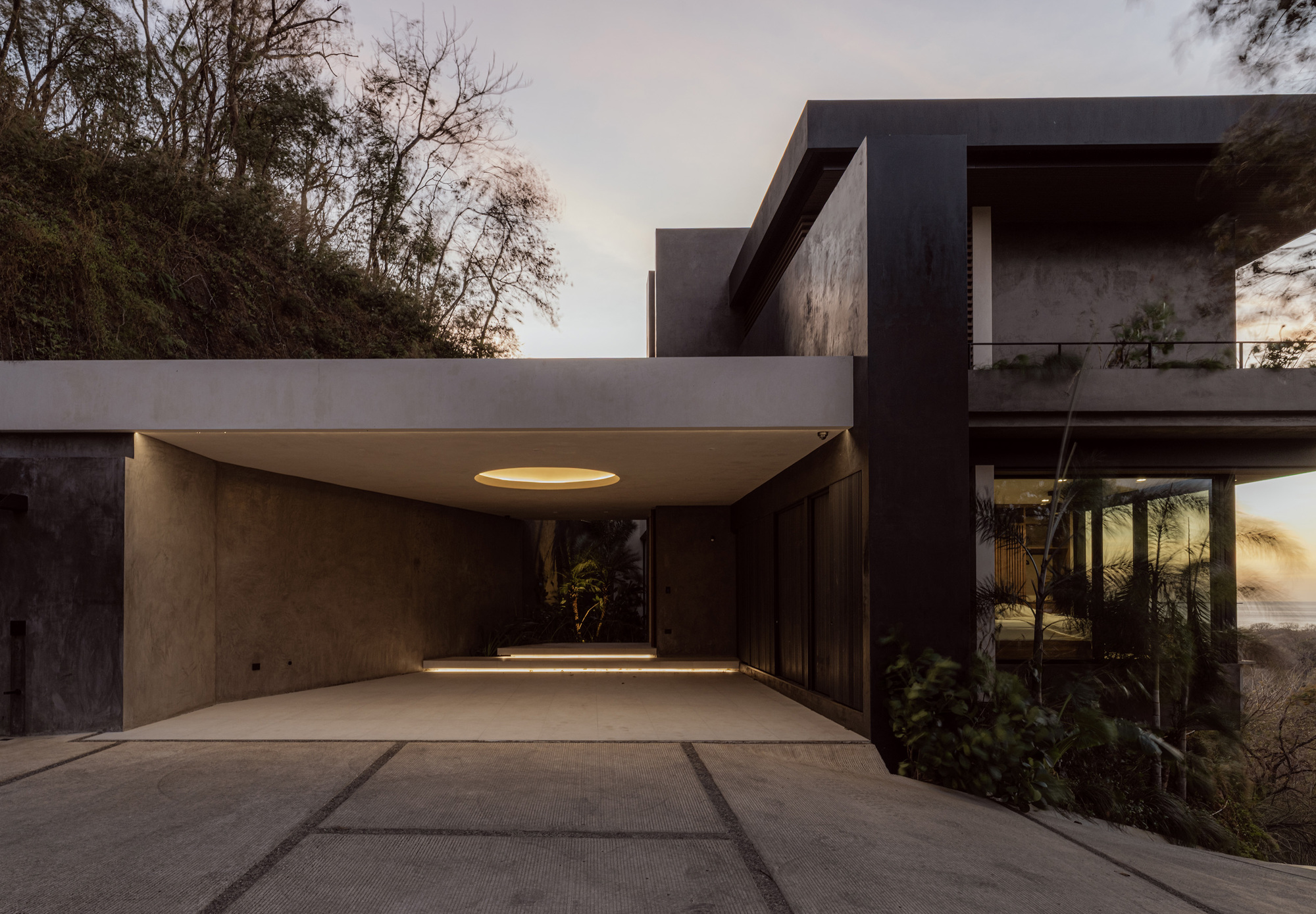 Concrete carport with a round skylight and dark stone walls, glowing at dusk beside a wooded slope