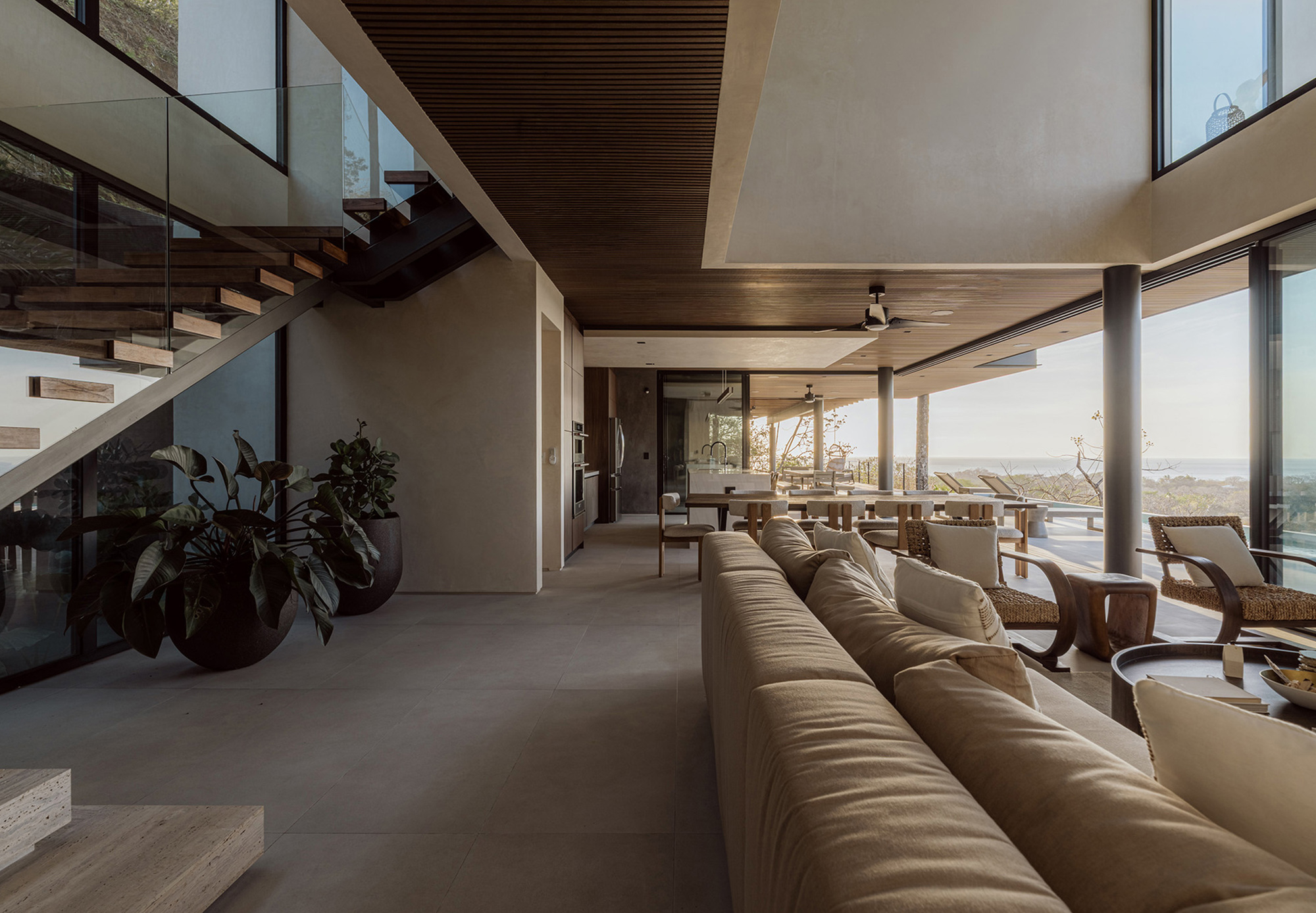 Double-height living space with a timber-tread staircase, long beige sofa and glazed wall opening toward a distant ocean horizon