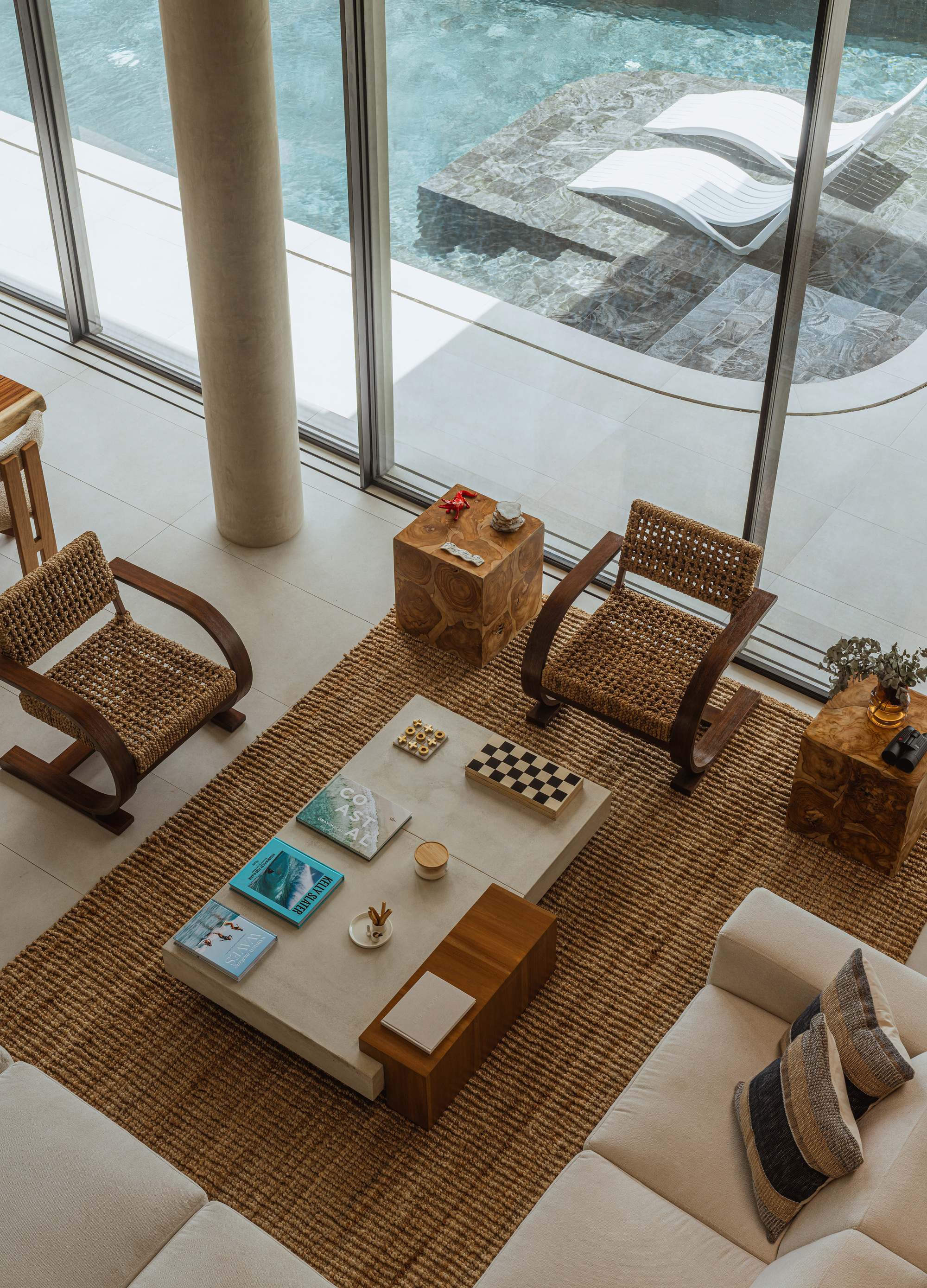 Top-down view of a living area with woven armchairs, a stone coffee table and a jute rug beside glazing onto the pool