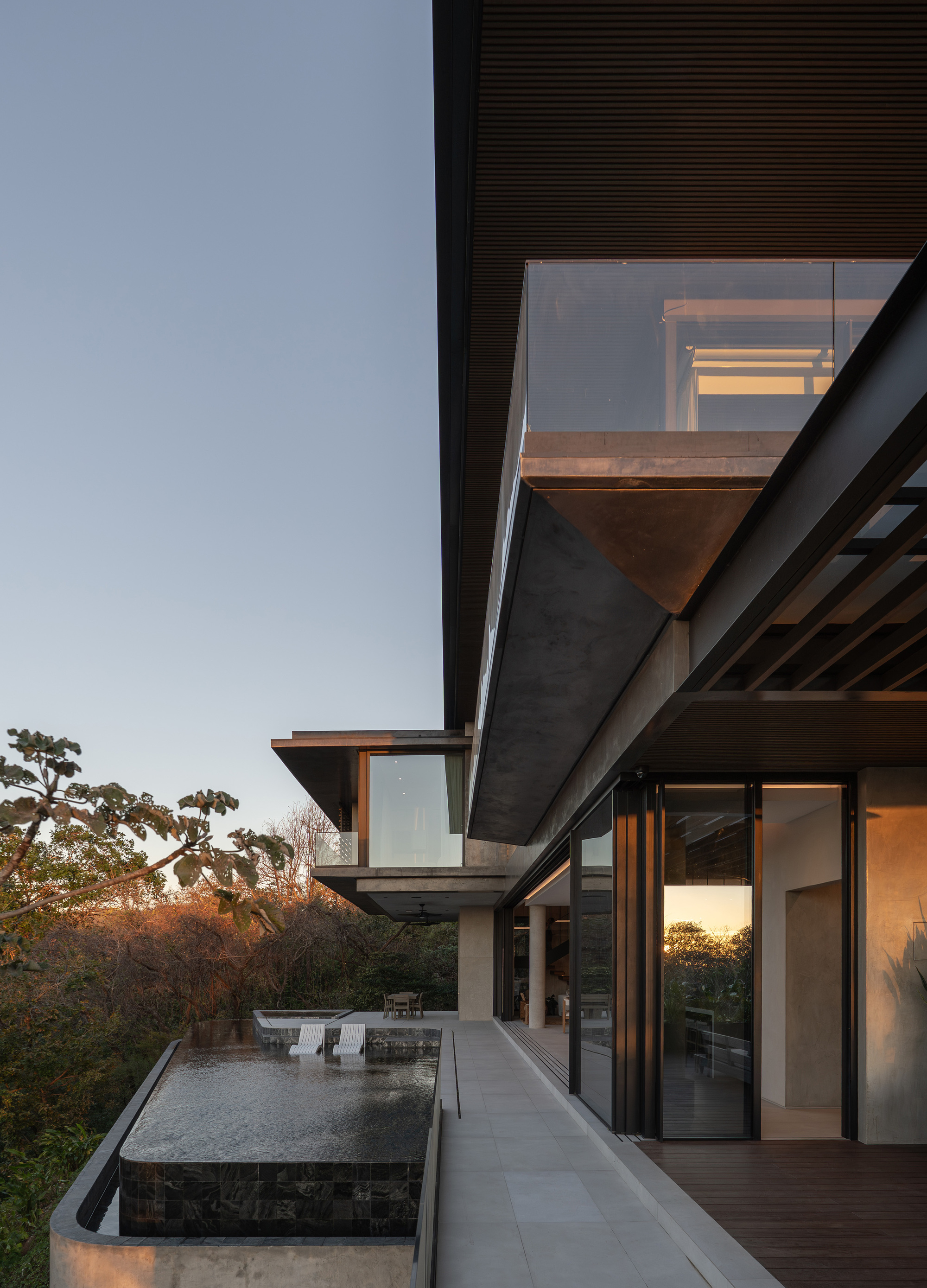 Cantilevered upper volume with glass balustrade above an infinity pool and terrace, lit by warm dusk light over treetops
